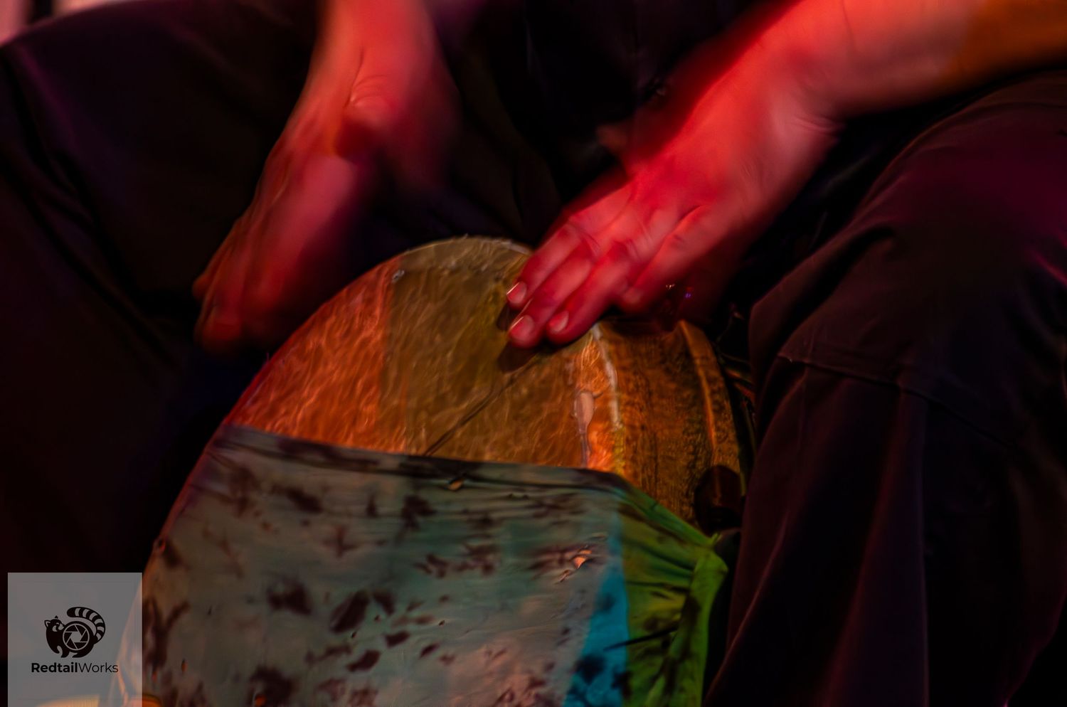 Close-up of hands playing a djembe drum under warm red stage lighting.