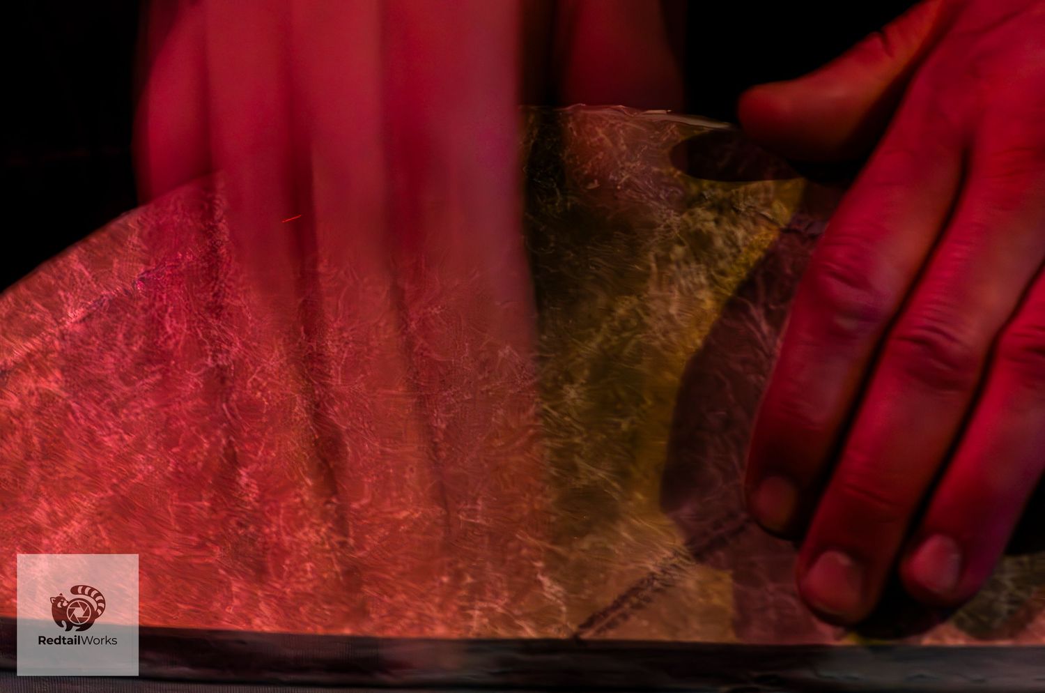 Abstract close-up of hands playing a cymbal bathed in red and yellow stage lighting.