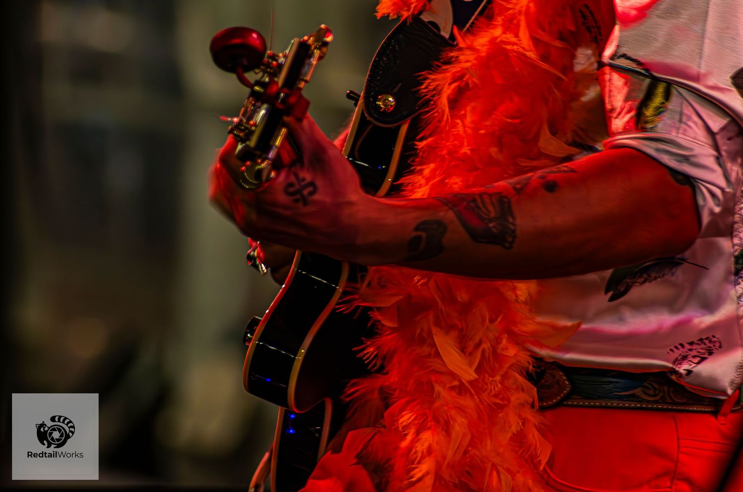 Guitarist with tattoos and red feather boa playing electric guitar on stage.