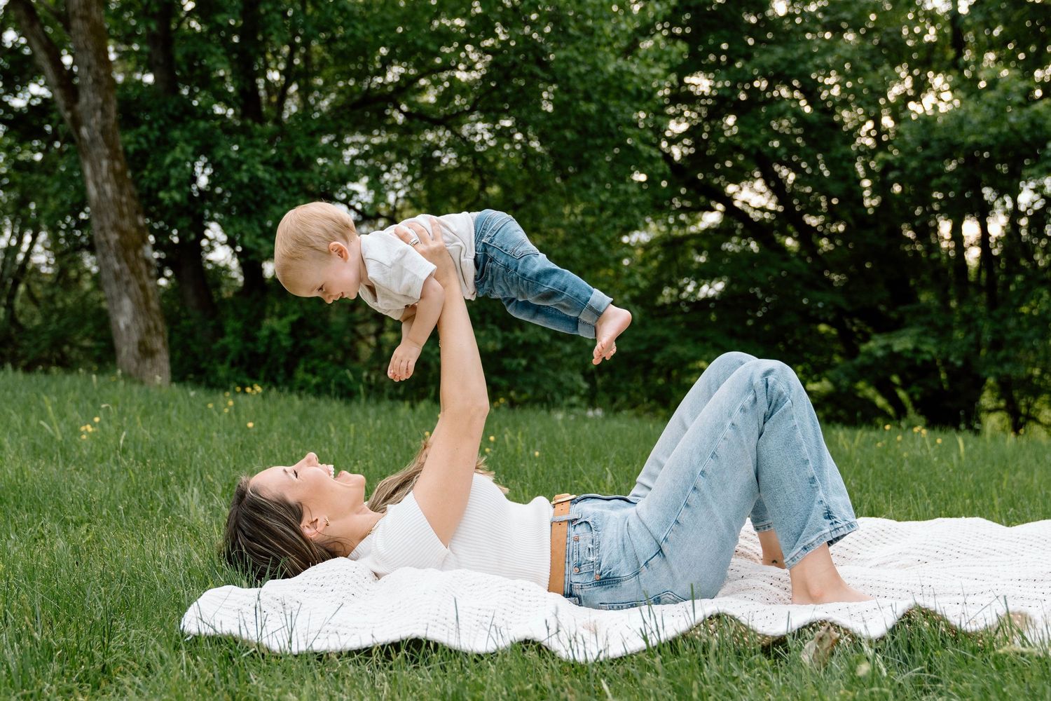 A mom lying on a picnic blanket holding her son up in the air while smiling