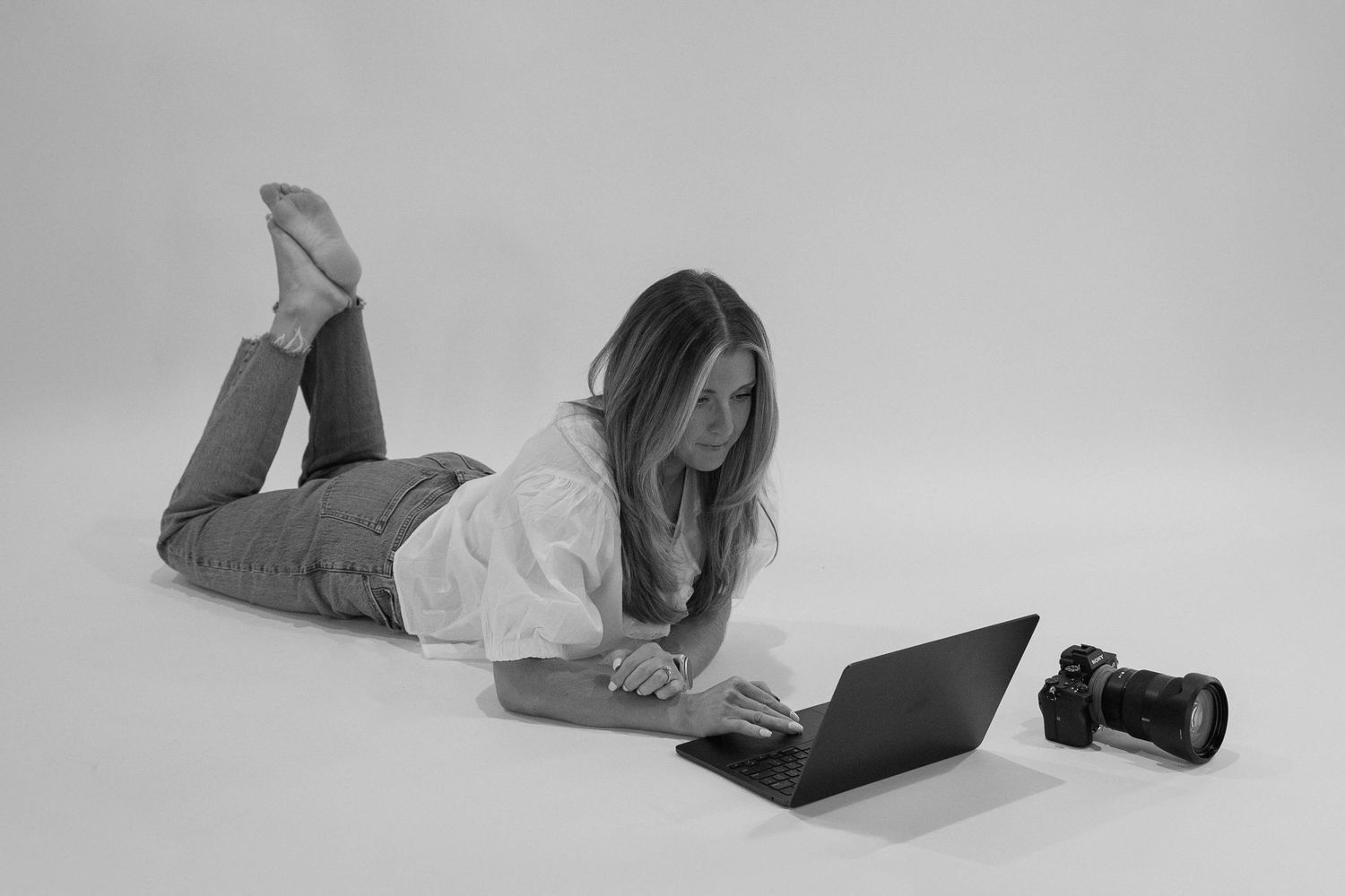 A woman lying on her stomach propped up on her elbows with her laptop and camera