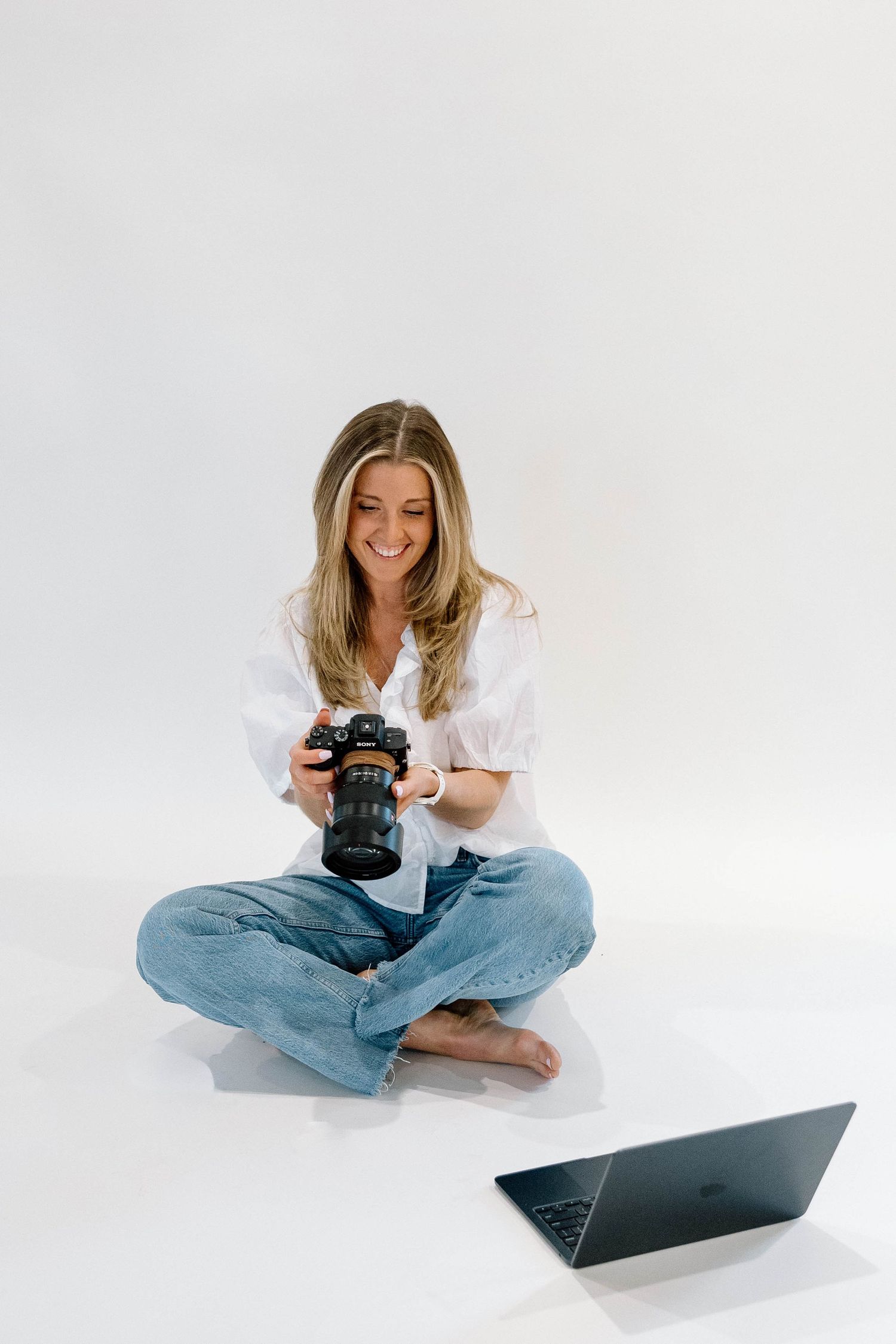 A woman sitting criss-crossed, looking at her camera with a laptop next to her