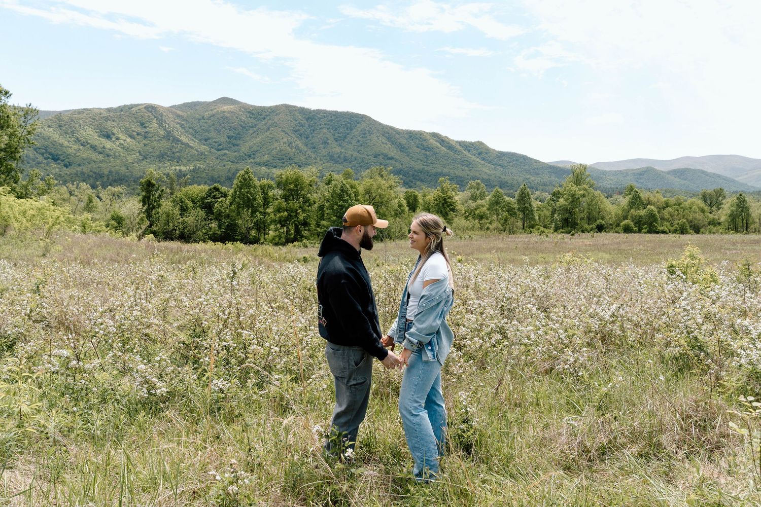A couple holding hands and facing each other in a field with mountains behind them
