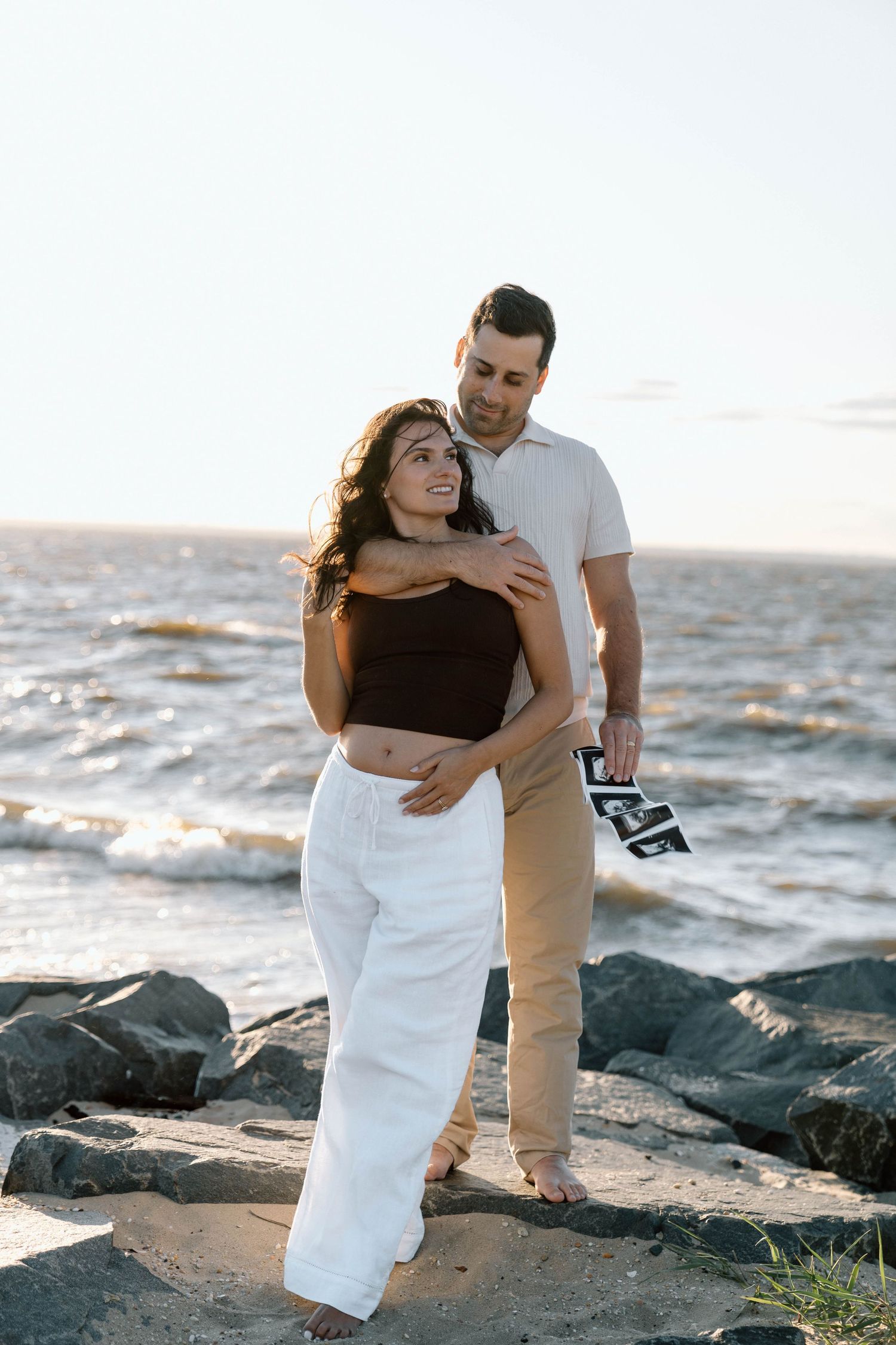 Husband hugging wife from behind on a beach with ultrasound images in his hand