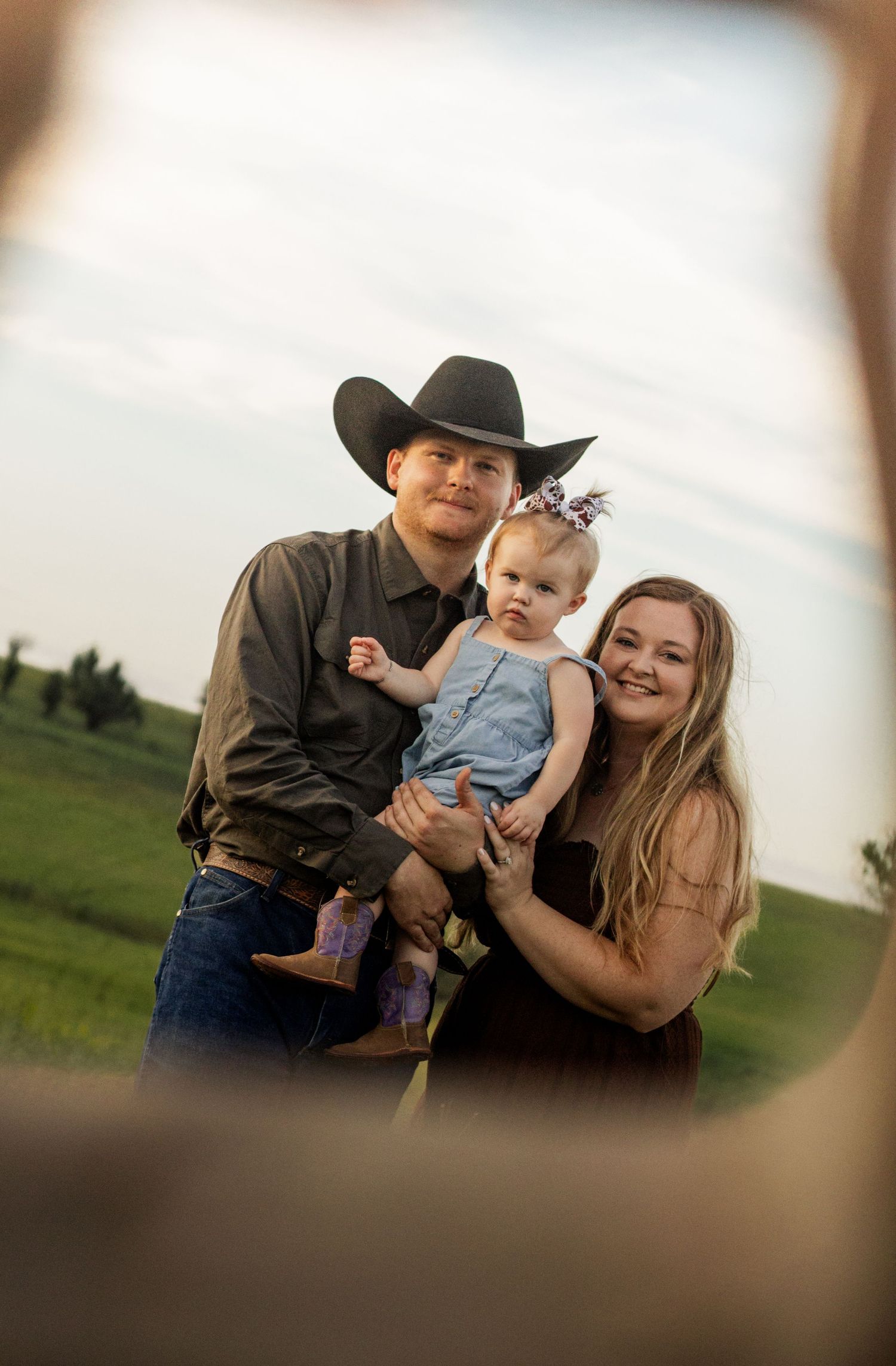 A young family with a man in a black cowboy hat holding a baby girl with a bow, standing with a smiling woman in a green field.