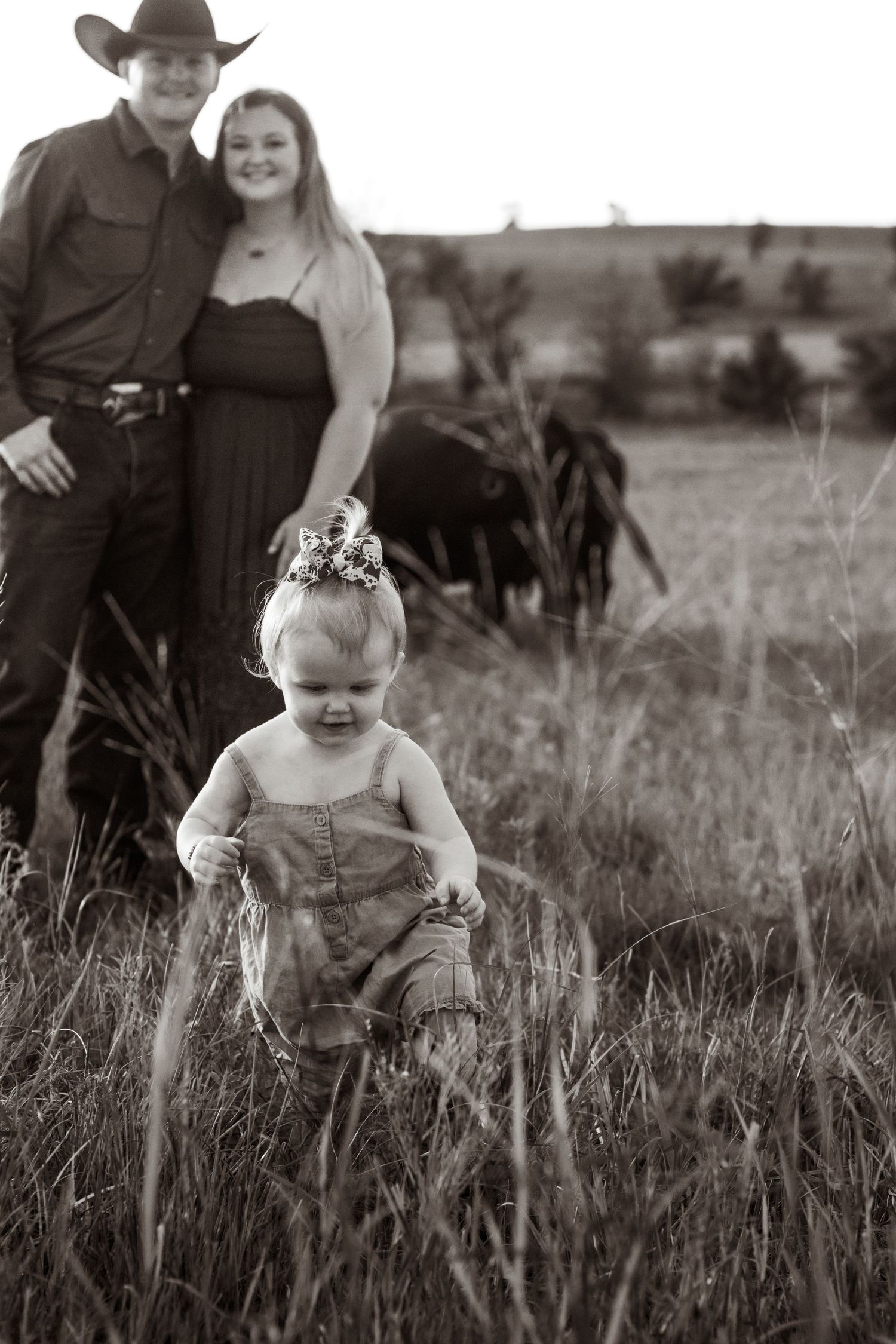 Black and white family photo of couple and toddler girl in flower crown in grassy field with cattle grazing behind them.
