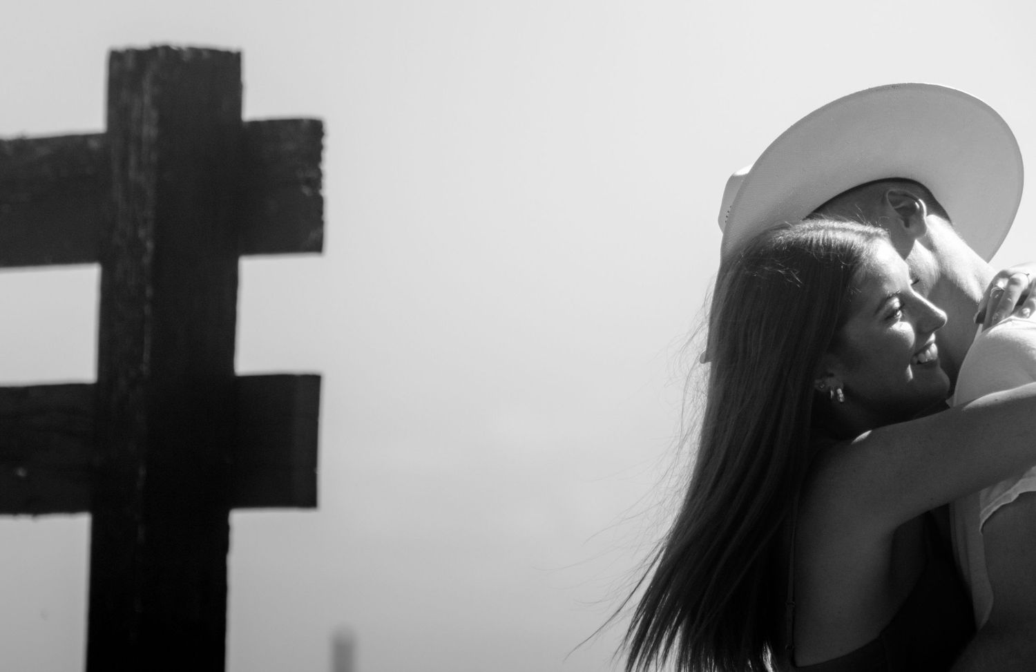 Black and white photo of a couple sharing a romantic moment near a large cross outdoors.