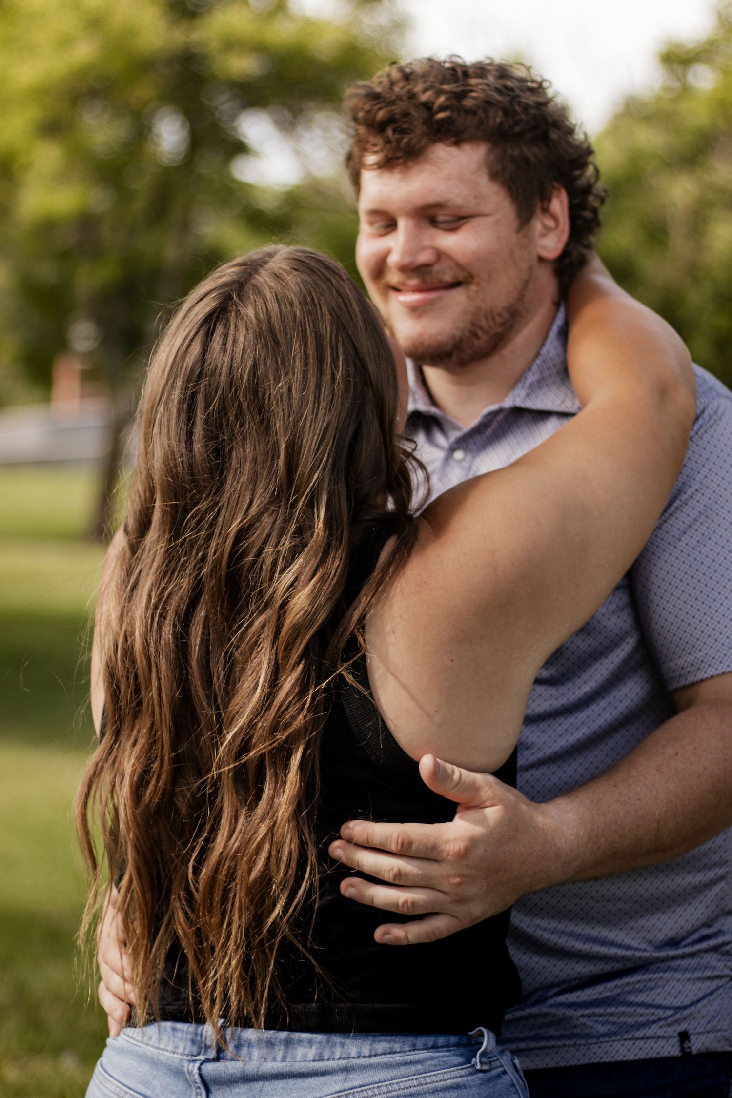 A couple shares a warm embrace outdoors, the man smiling as he holds a woman with long brown hair in a park.