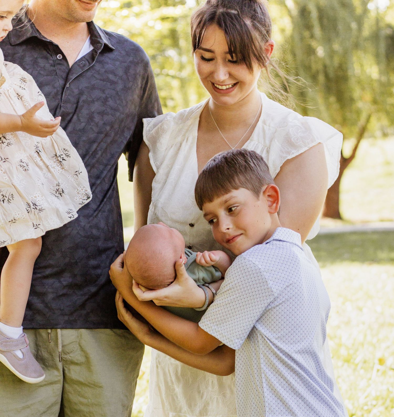 Family of four outdoors, parents holding newborn baby and toddler, older boy smiling, surrounded by green trees.