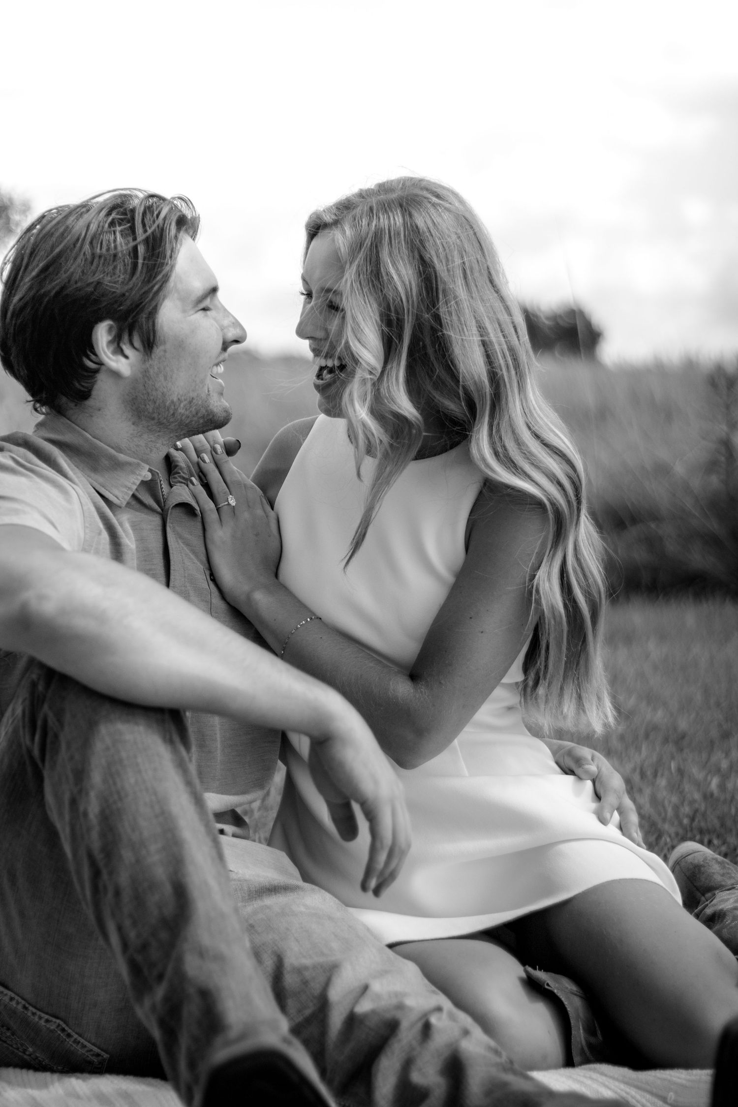 Couple sitting outdoors laughing and gazing at each other in a black and white engagement photo.