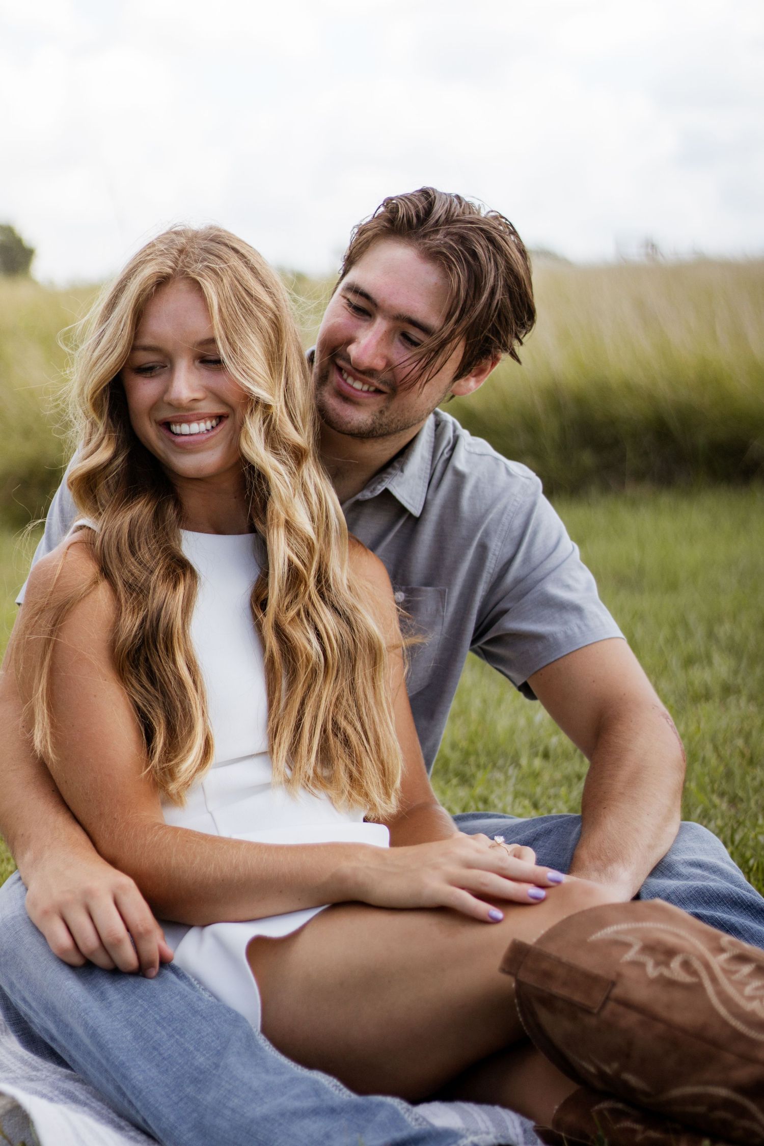 A smiling couple sits together in a grassy field, the woman with long blonde hair leaning against the man.