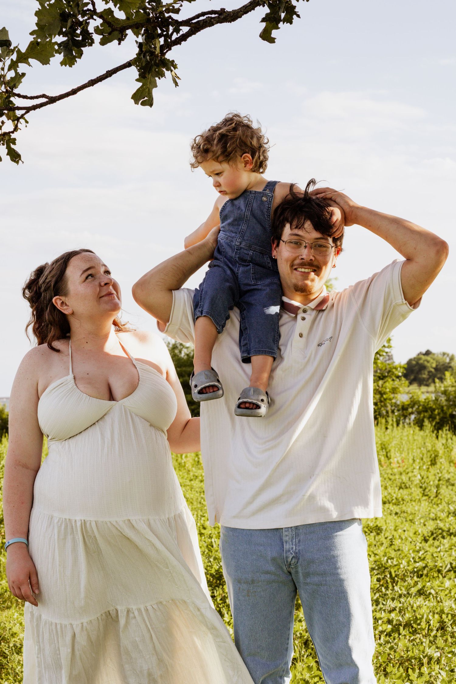 Pregnant woman in white dress stands with man carrying toddler on shoulders in sunny green field.