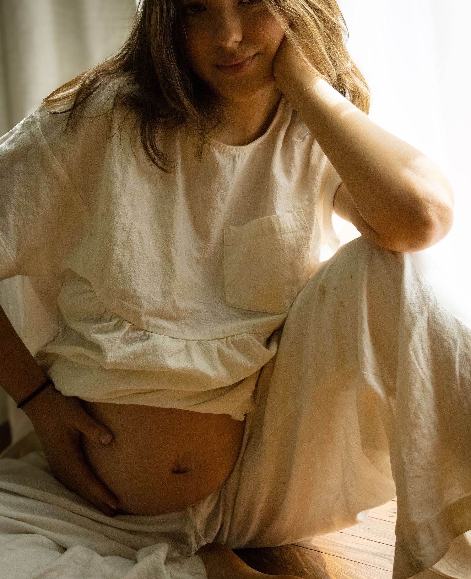 Pregnant woman in white linen outfit sitting on floor gently holding her bare baby bump near window.