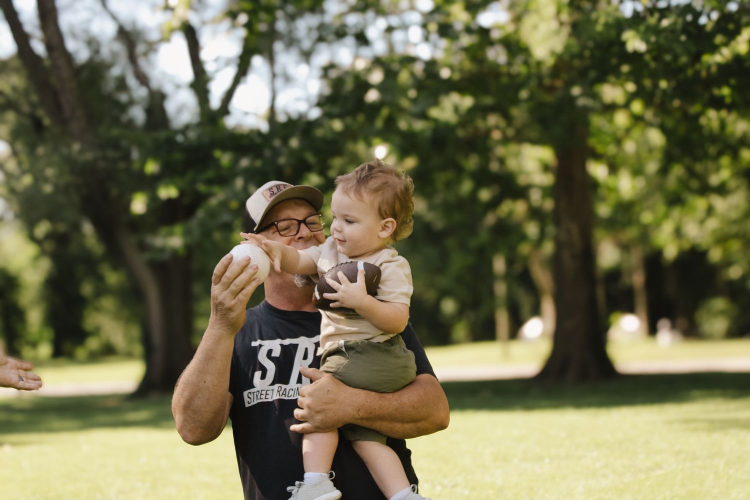 Man in SRC cap and black t-shirt playing with a curious toddler outdoors in a green park.