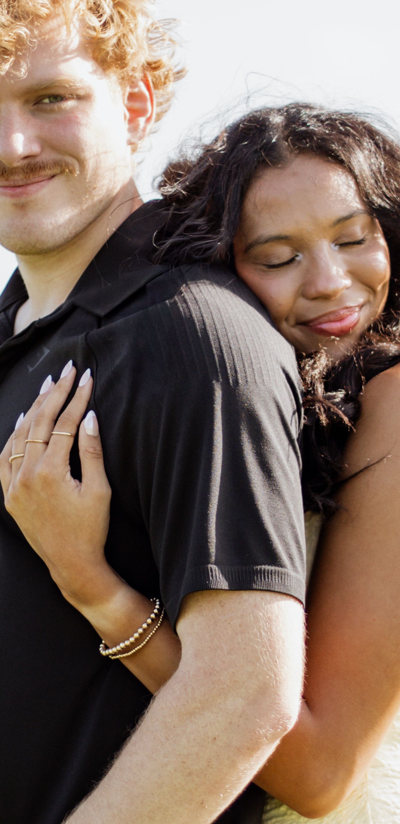 A joyful interracial couple embracing outdoors, woman hugging man from behind, both smiling warmly.