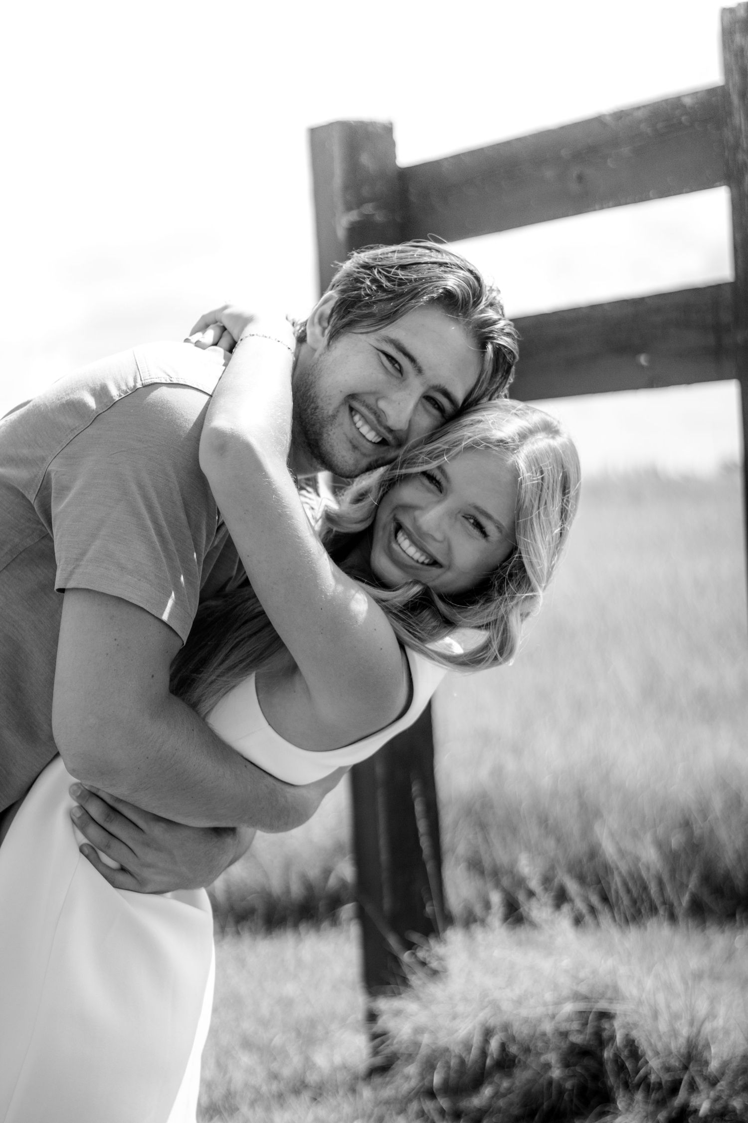 Happy couple hugging and laughing in black and white photo near wooden fence in grassy field.