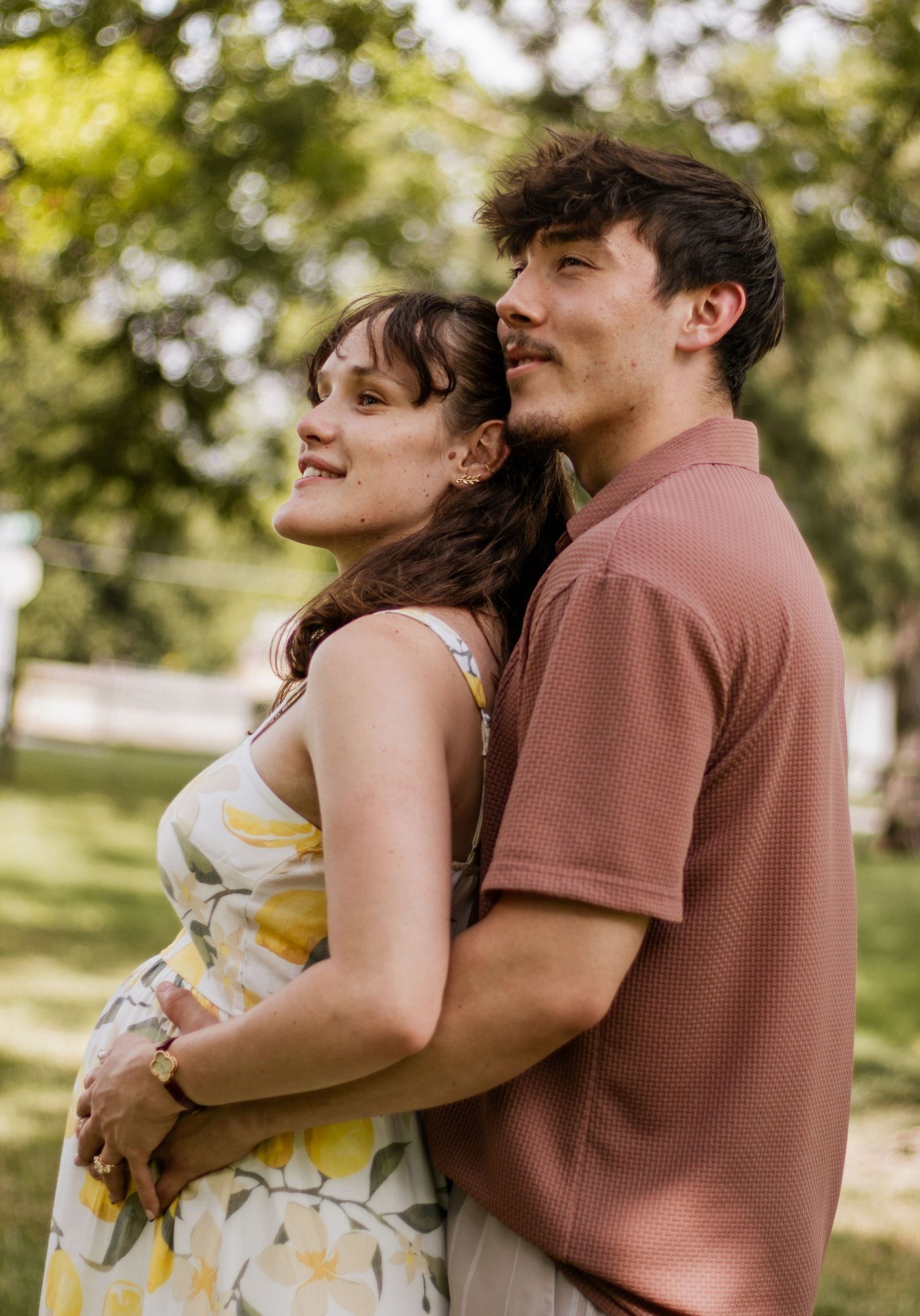 A couple embraces in a sunny park, the woman in a floral dress cradling her pregnant belly.