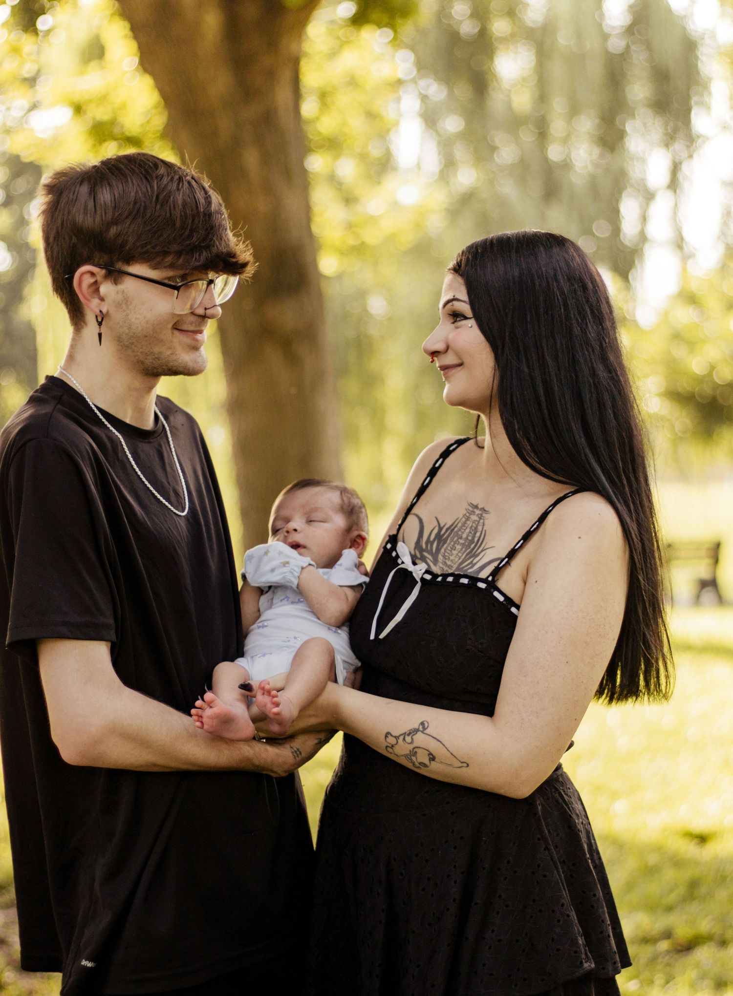 Young couple in black outfits holding newborn baby outdoors in sunny park setting.