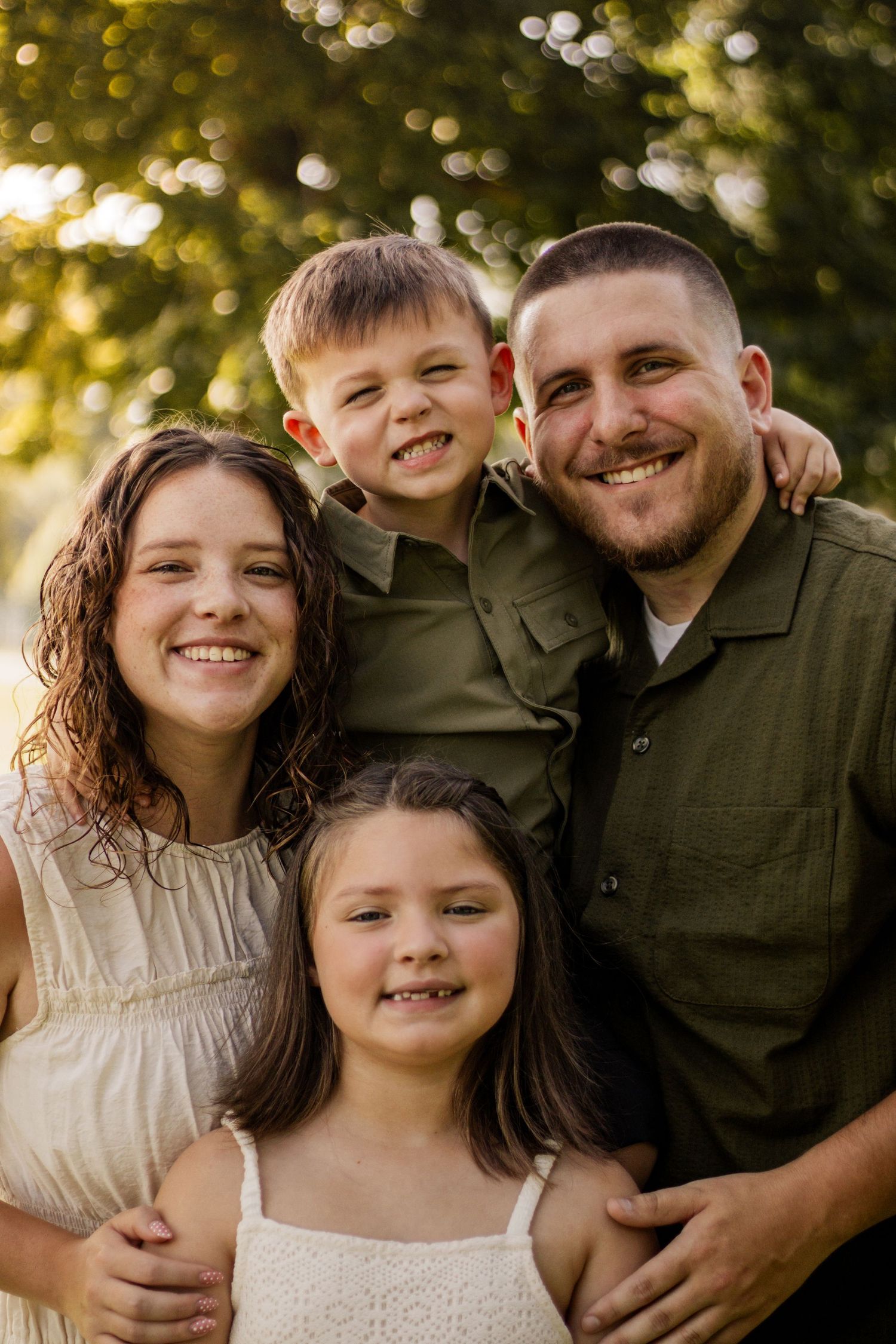 Joyful family of four smiling closely together outdoors with warm bokeh lighting in background.
