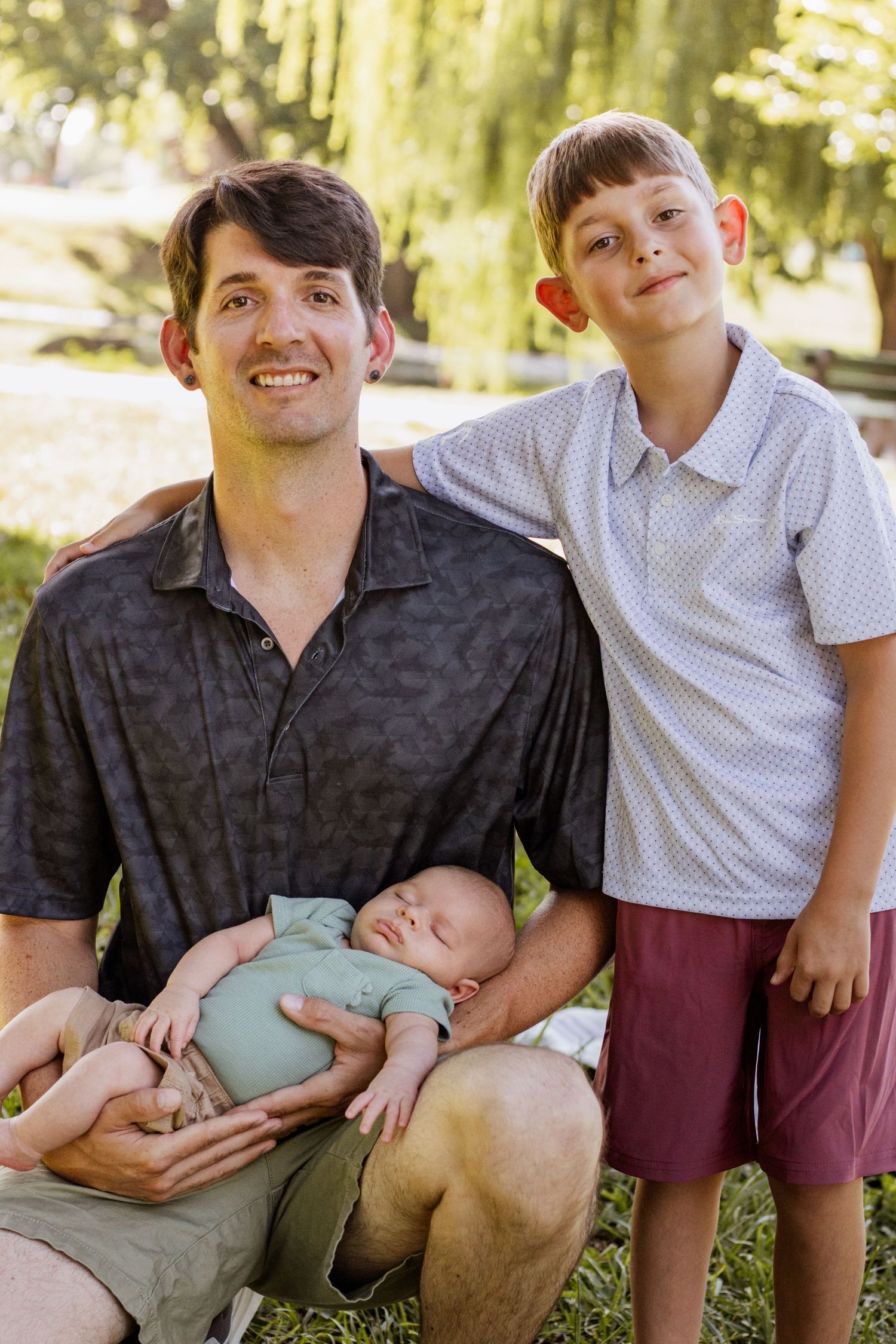 Father holding sleeping baby outdoors in park with older son standing beside him, smiling family portrait.