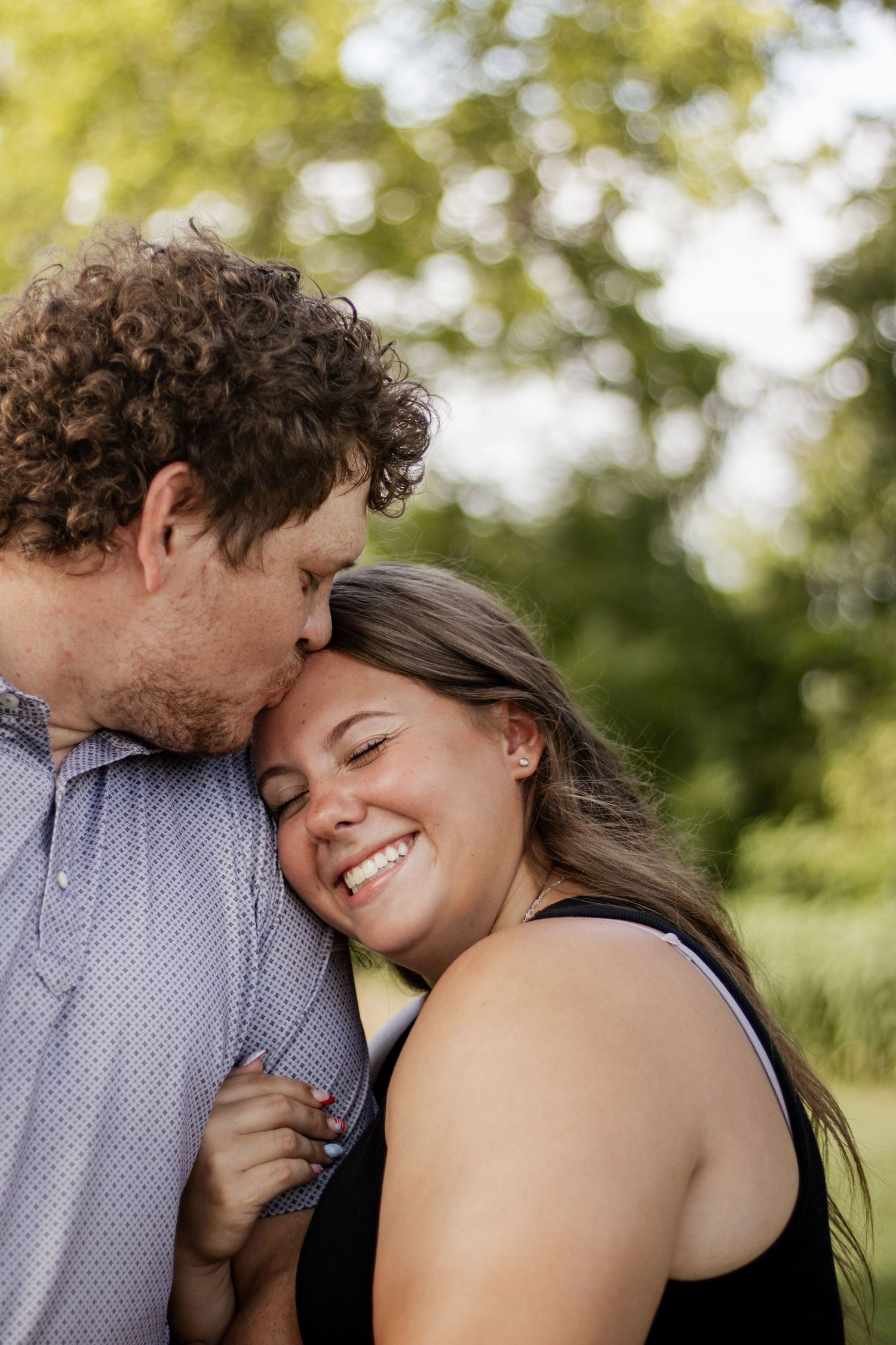 A man with curly hair kisses a smiling woman on the cheek outdoors, surrounded by lush green trees.