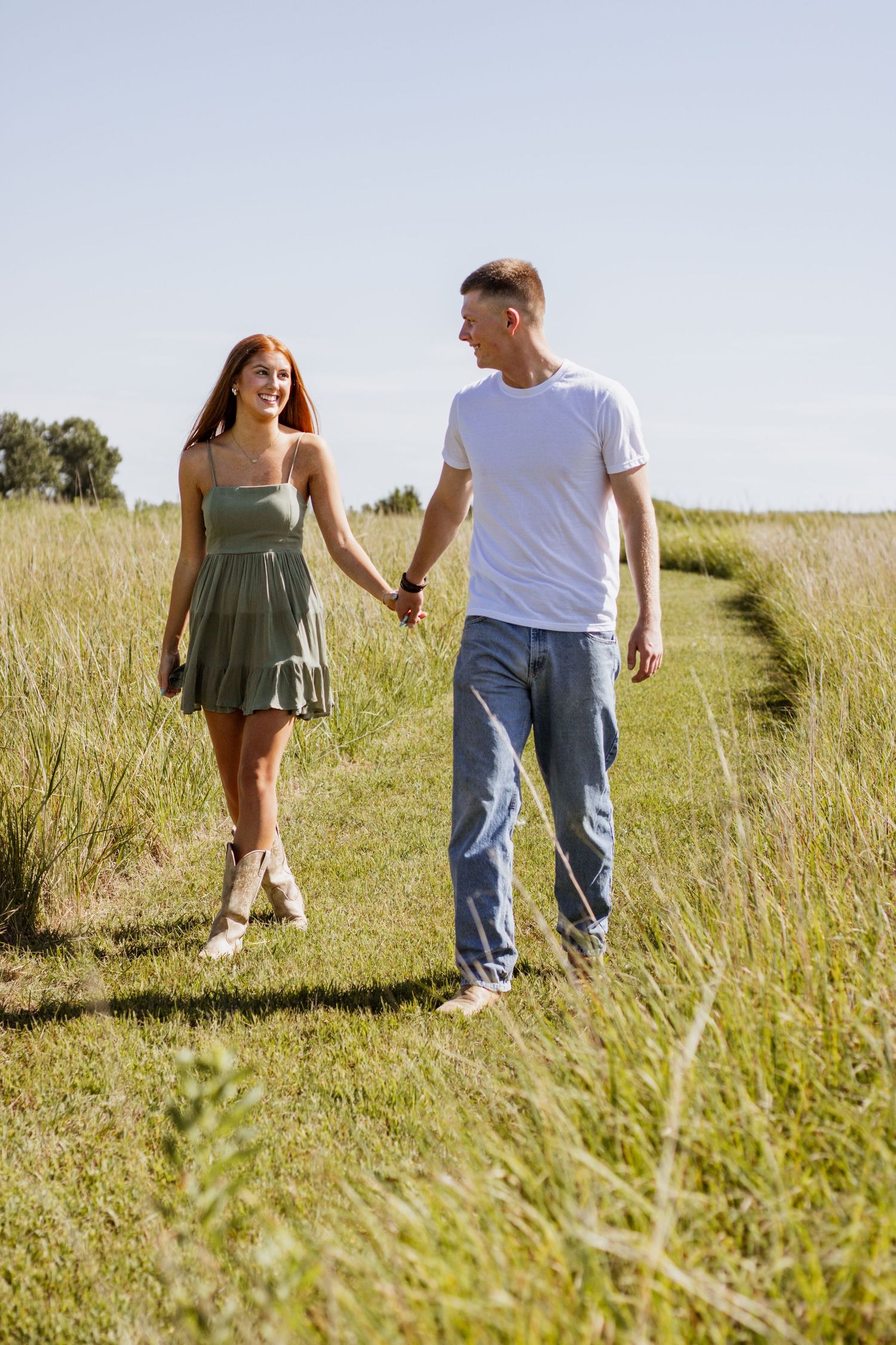 Young couple holding hands walking through a sunny green field, woman in olive dress, man in white t-shirt and jeans.