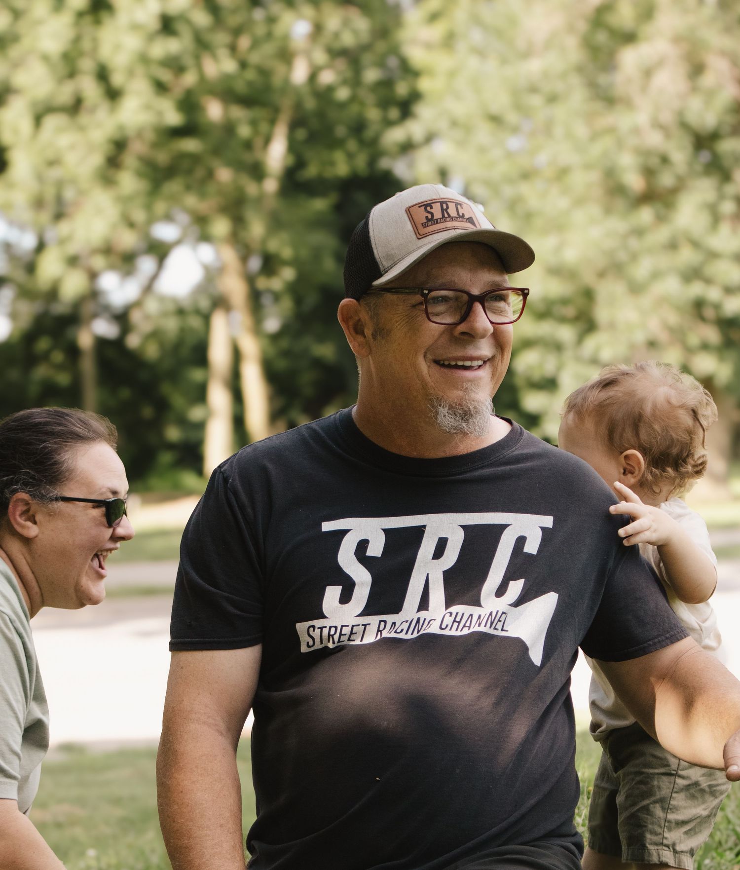 Smiling man in SRC Street Racing Channel t-shirt laughing outdoors in a sunny green park.
