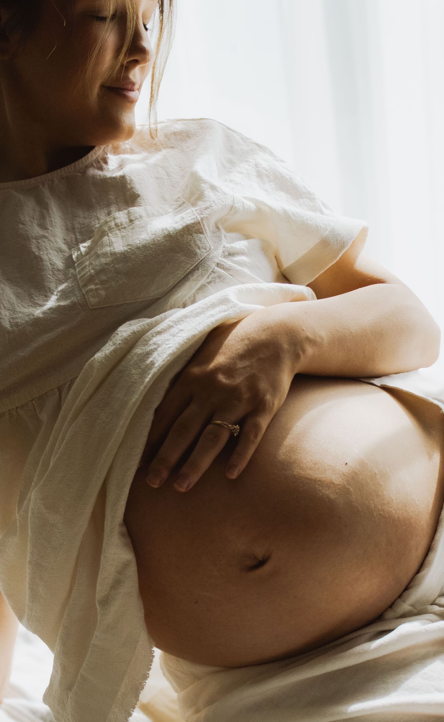 Pregnant woman in white linen dress cradling her bare belly with one hand wearing a ring near window.