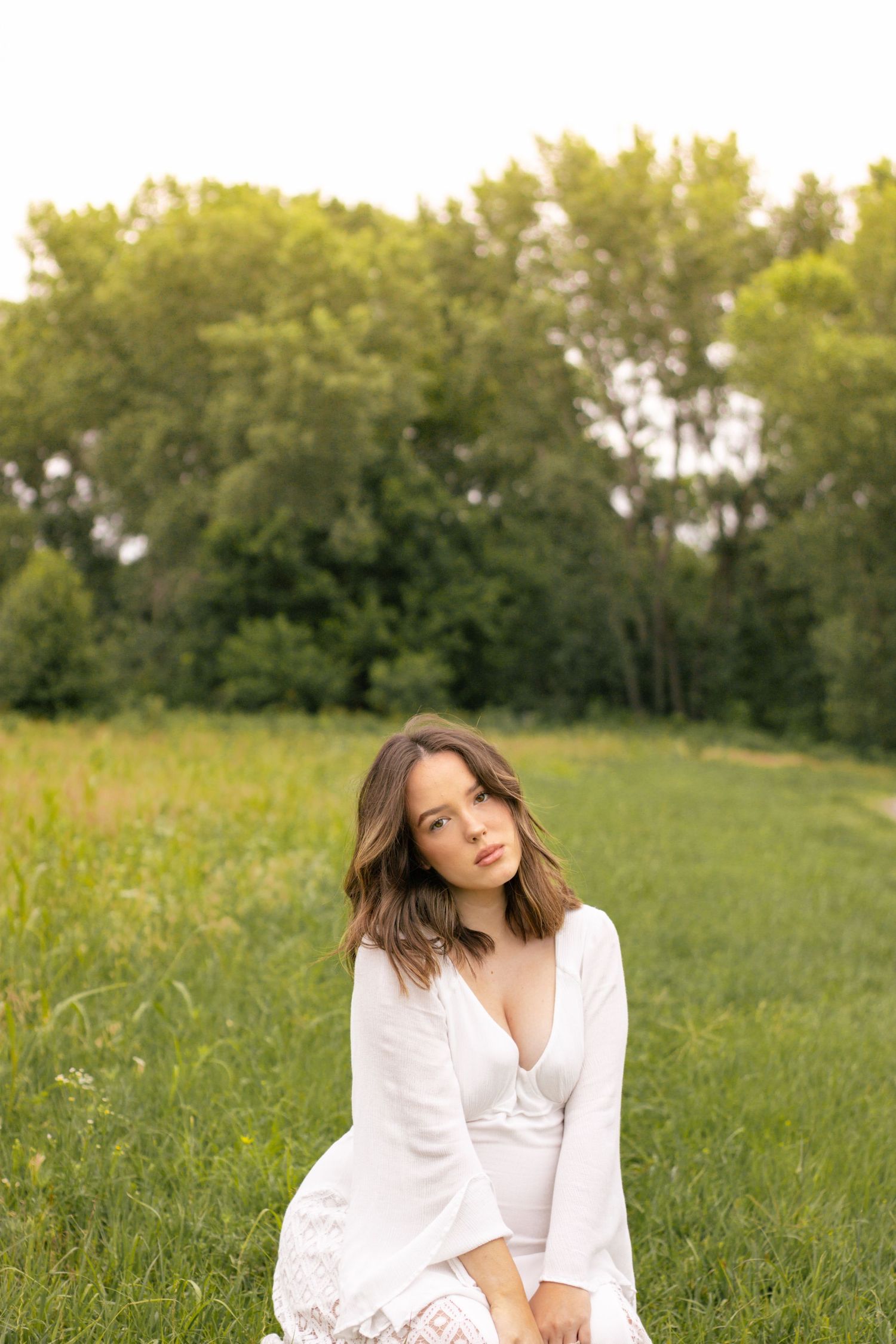 Young brunette woman in white dress sitting in green grassy field with trees background.