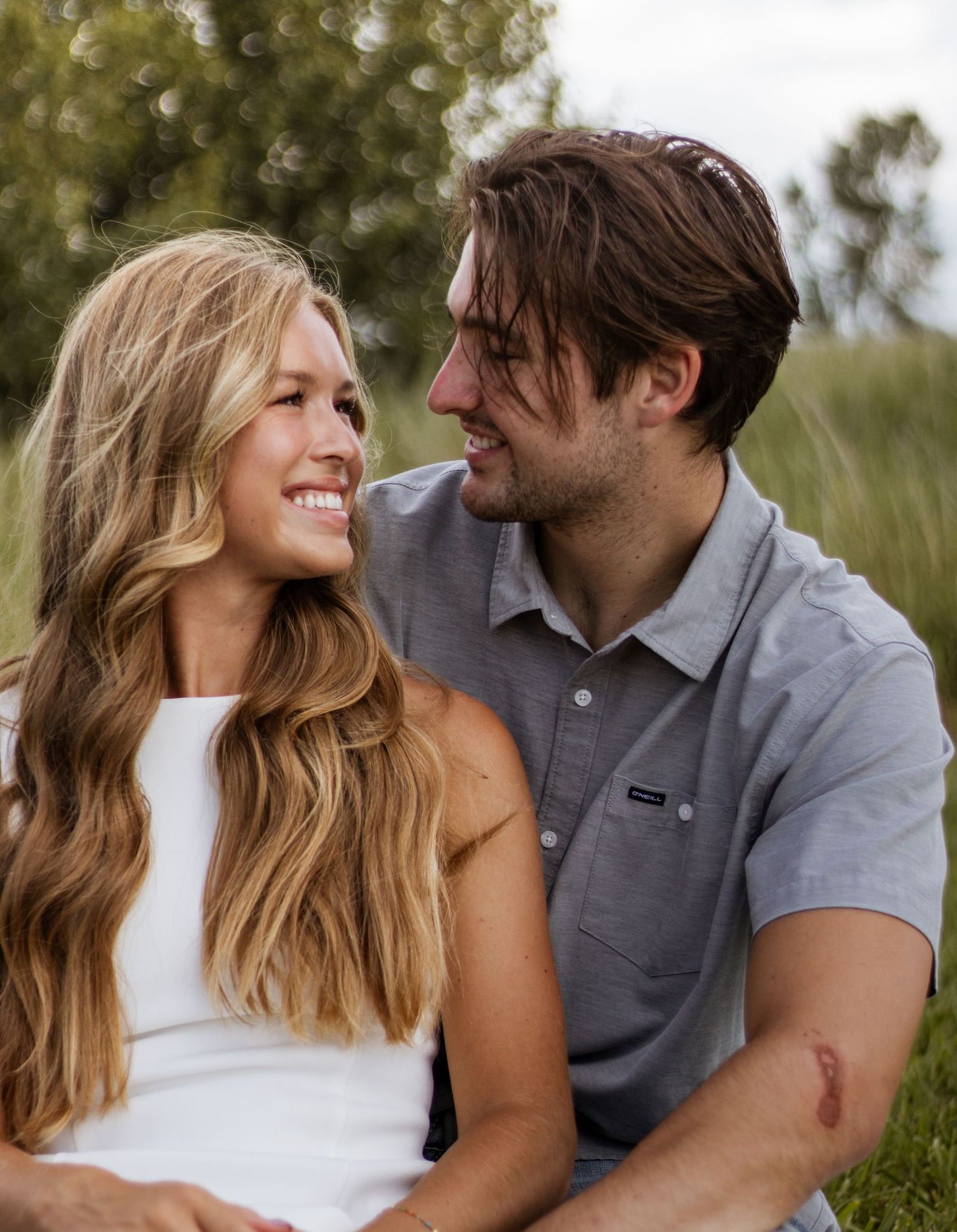 A smiling young couple sitting outdoors in a green field, gazing at each other affectionately on a sunny day.