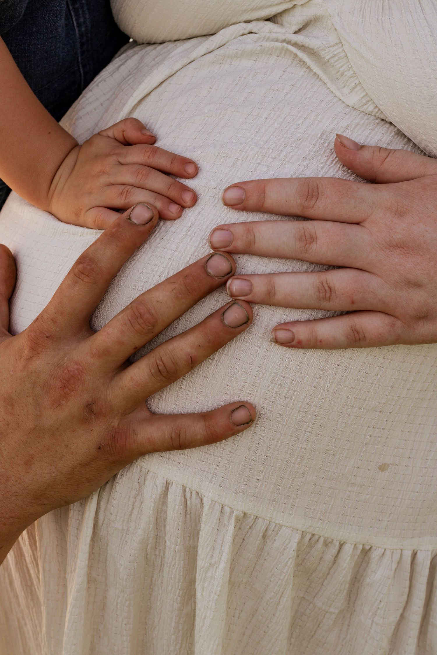 Close-up of pregnant woman's belly with partner and child's hands gently resting on white dress.