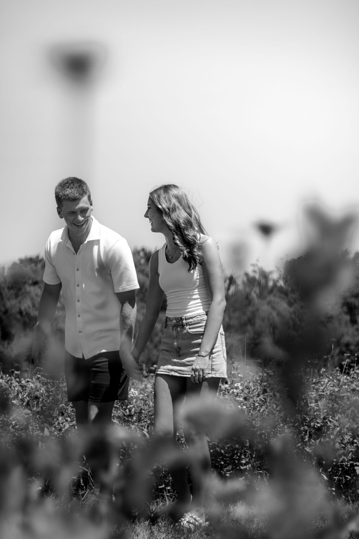 Black and white photo of a young couple holding hands and smiling while walking through a lush outdoor field.
