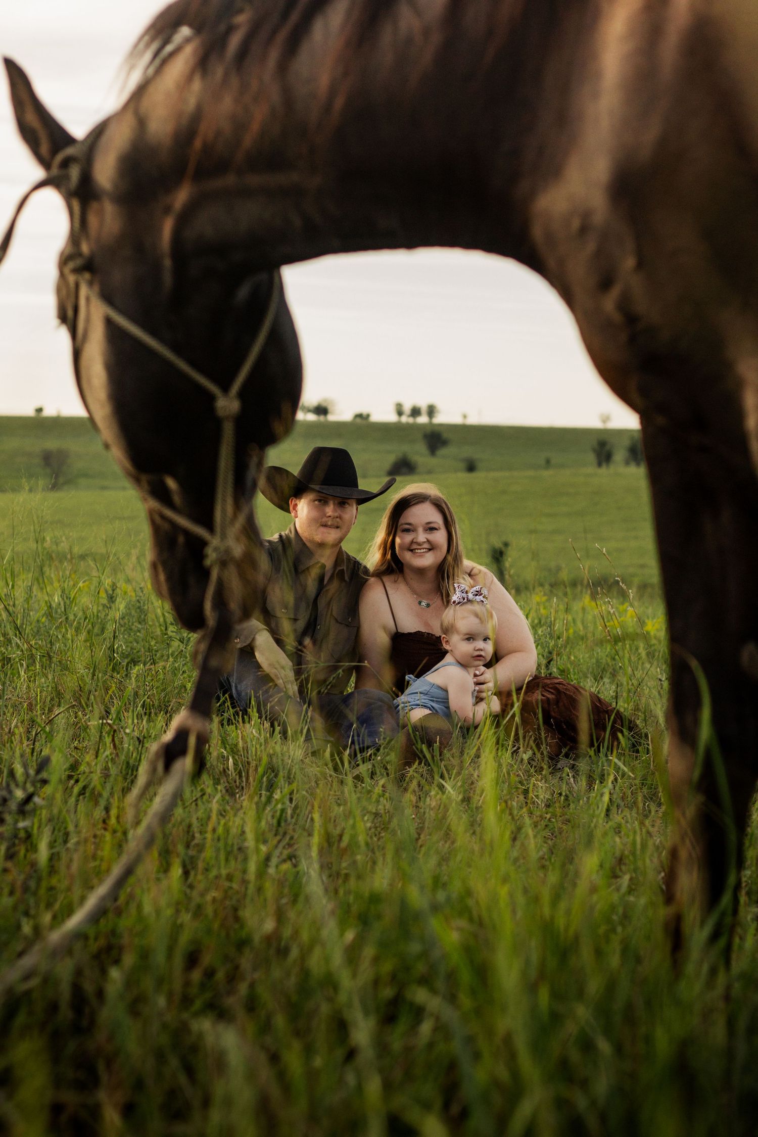 Family of three sitting in green field with dark horse in foreground, man in cowboy hat, woman holding baby, rural setting.