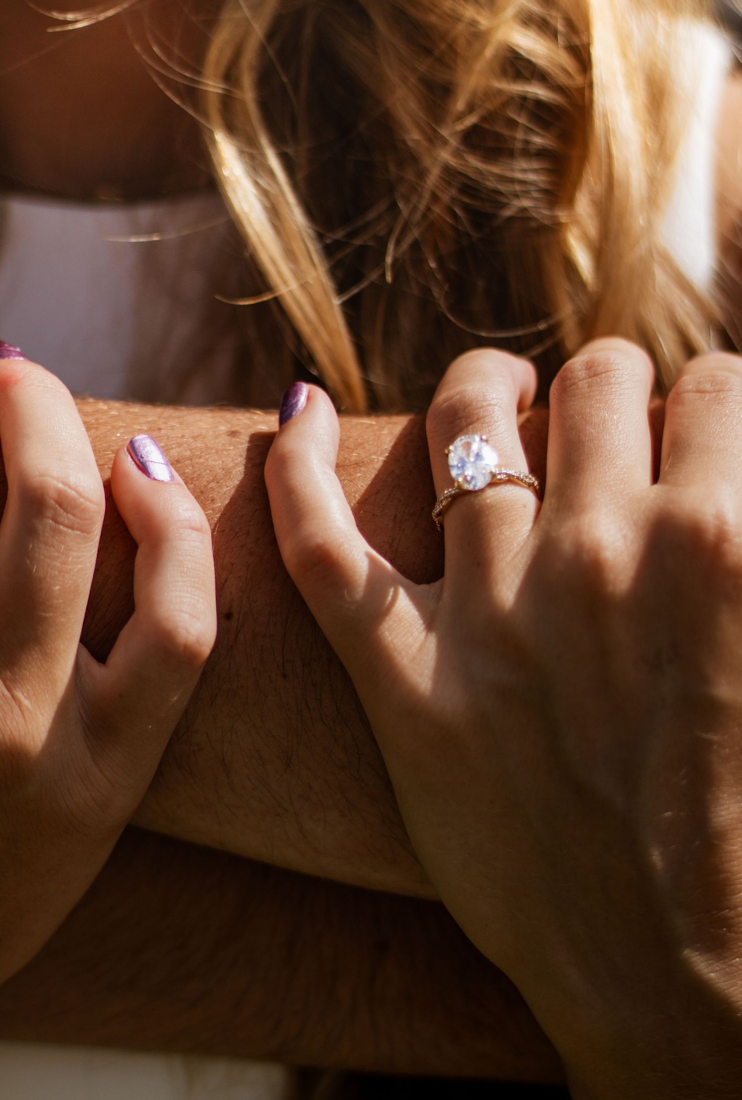 Close-up of hands resting on wood, one wearing a large oval diamond engagement ring, with purple manicured nails visible.