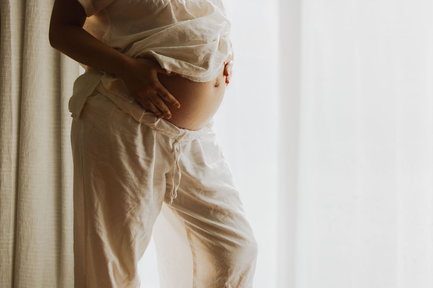 Side profile of pregnant woman in white outfit gently holding bare baby bump by bright window.
