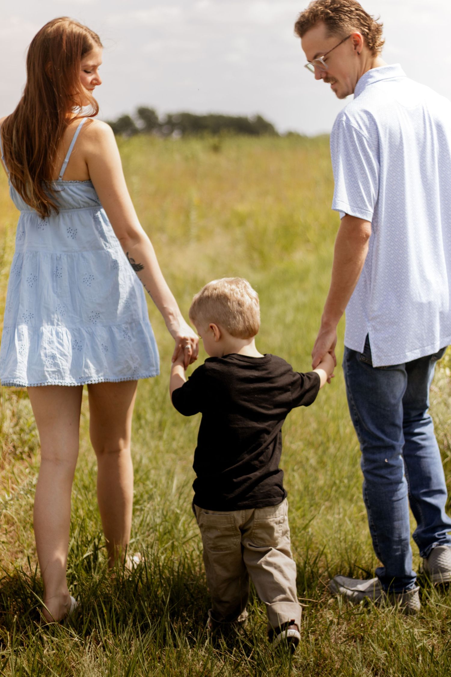 A young family with a toddler holding hands in a green field, parents in casual summer clothes enjoying outdoor time.