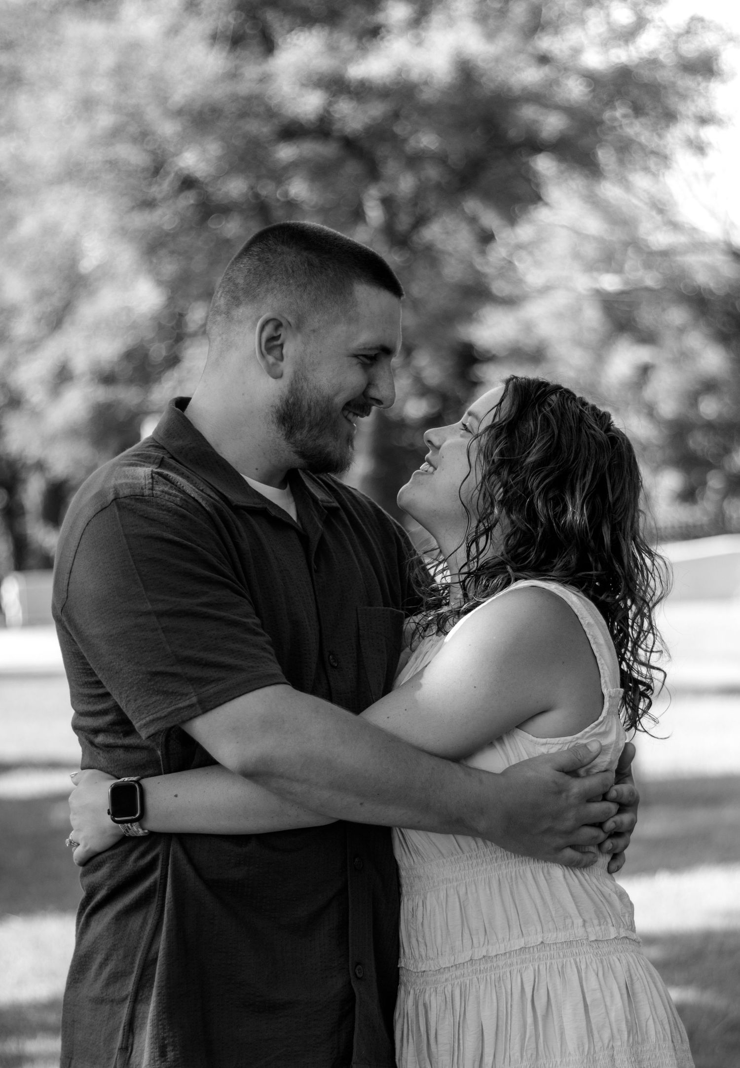 Black and white photo of a couple embracing and smiling at each other outdoors among trees.