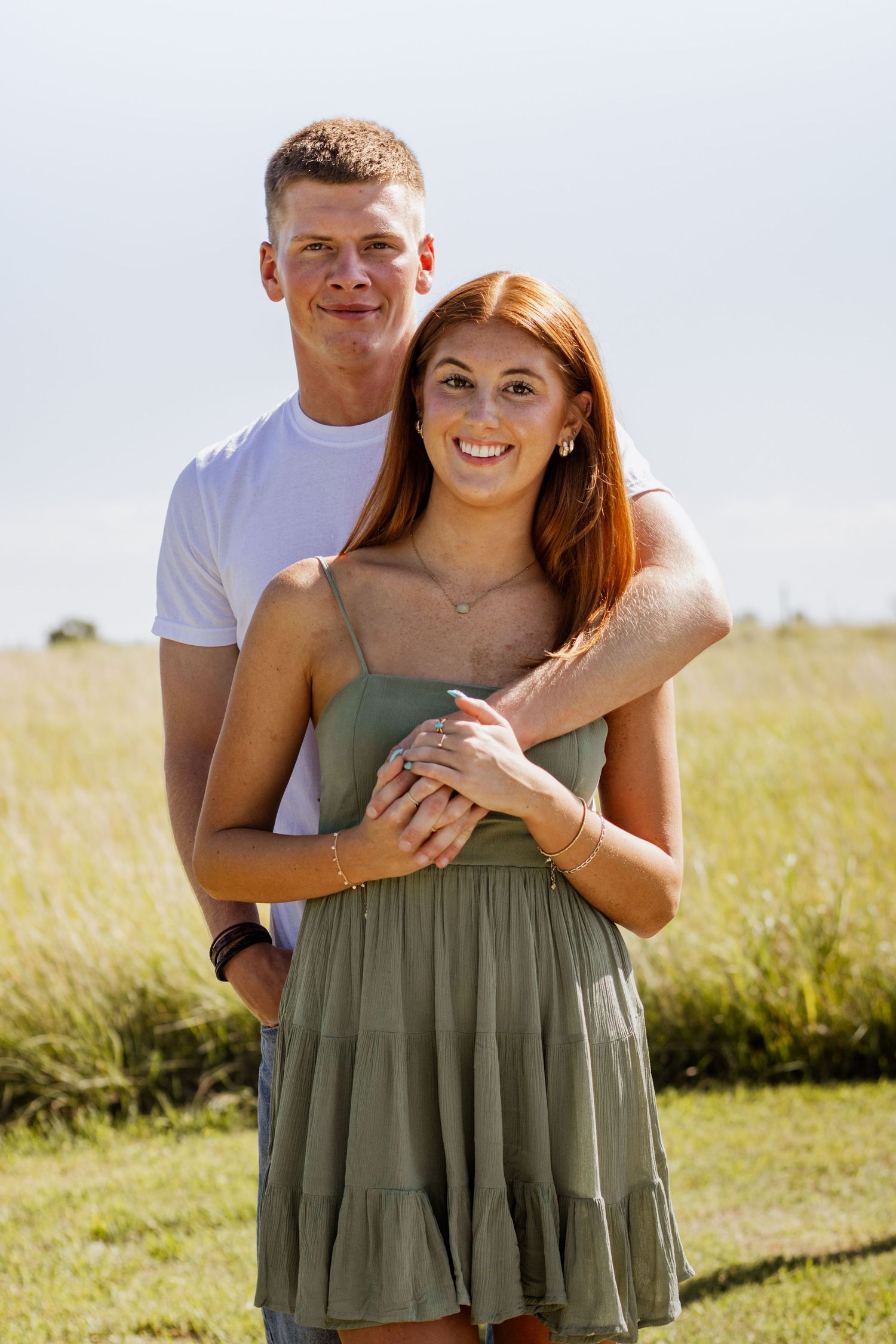 A smiling couple poses in a sunny field, the man in a white tee embracing a woman in a sage green sundress.