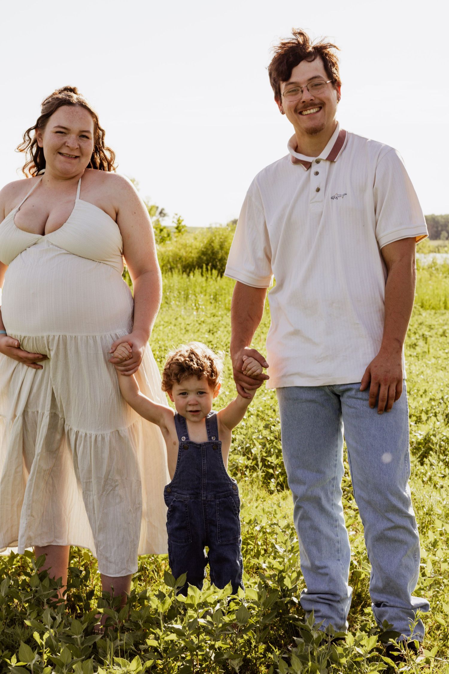 Pregnant woman in cream dress and man in white polo hold hands with toddler in overalls outdoors.