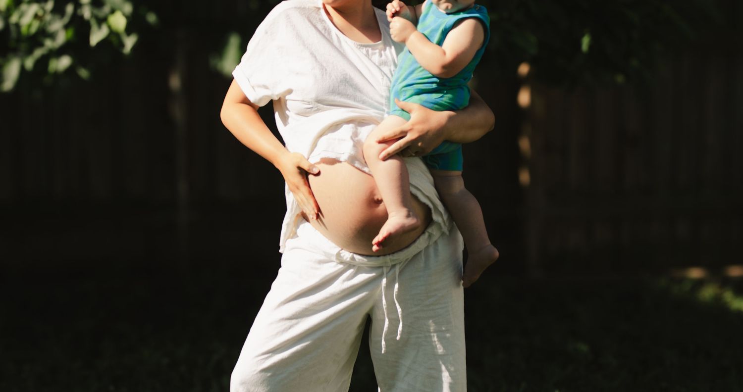 Pregnant woman in white outfit holds toddler in blue outdoors in sunny backyard setting.