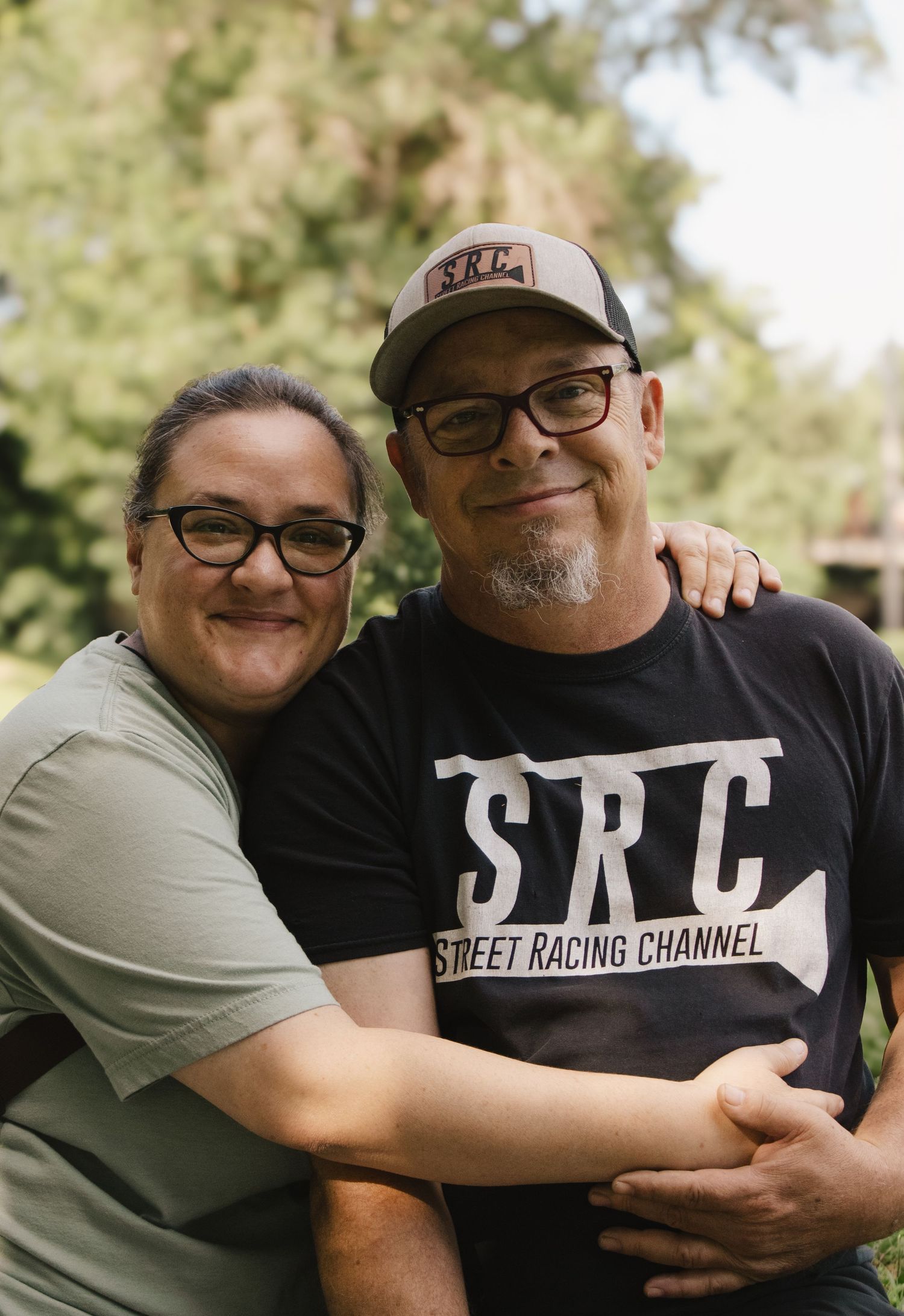 Two smiling men wearing glasses embrace outdoors, one in SRC Street Racing Channel black tee.