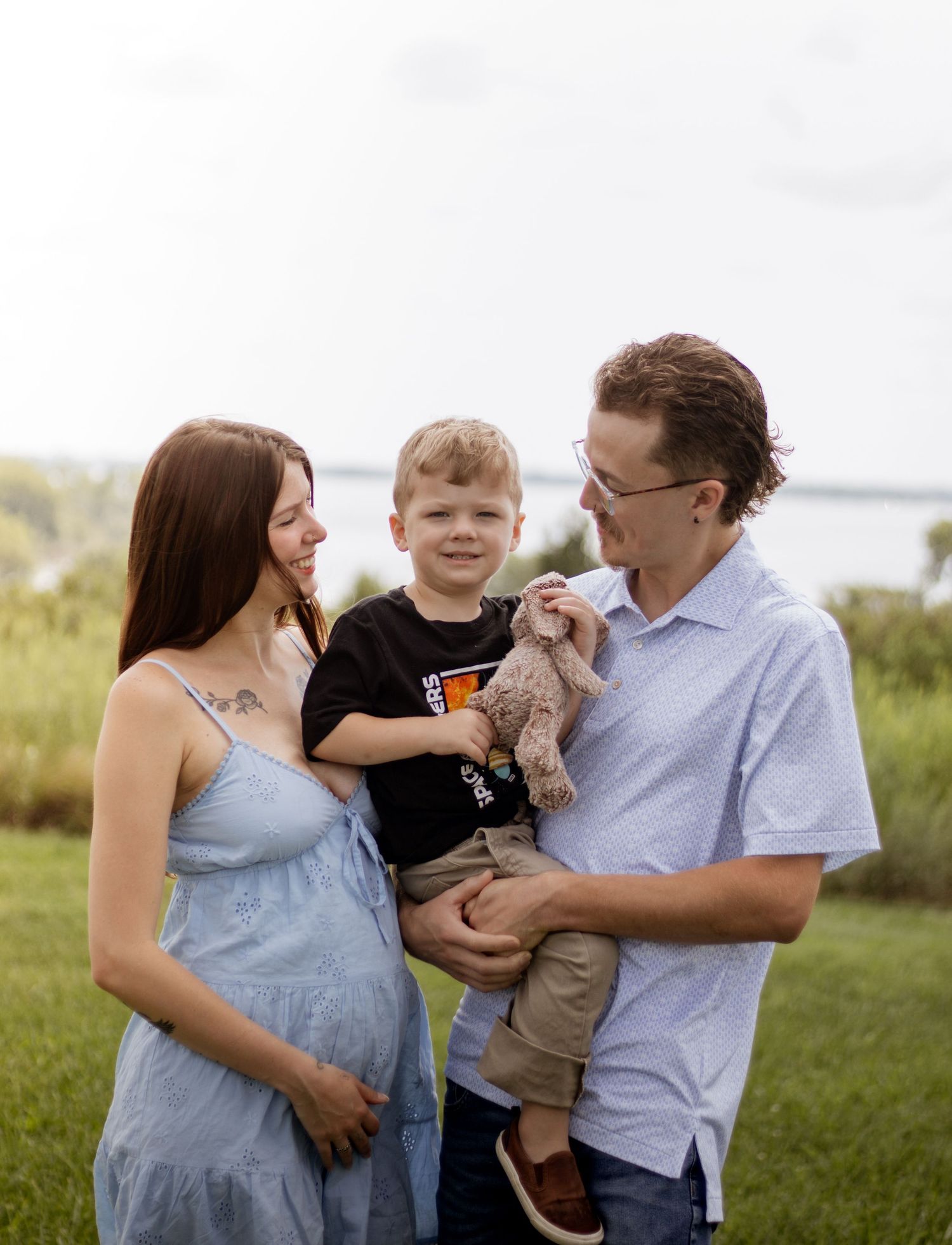 Pregnant woman in blue dress with husband holding toddler son outdoors near water on green grass.