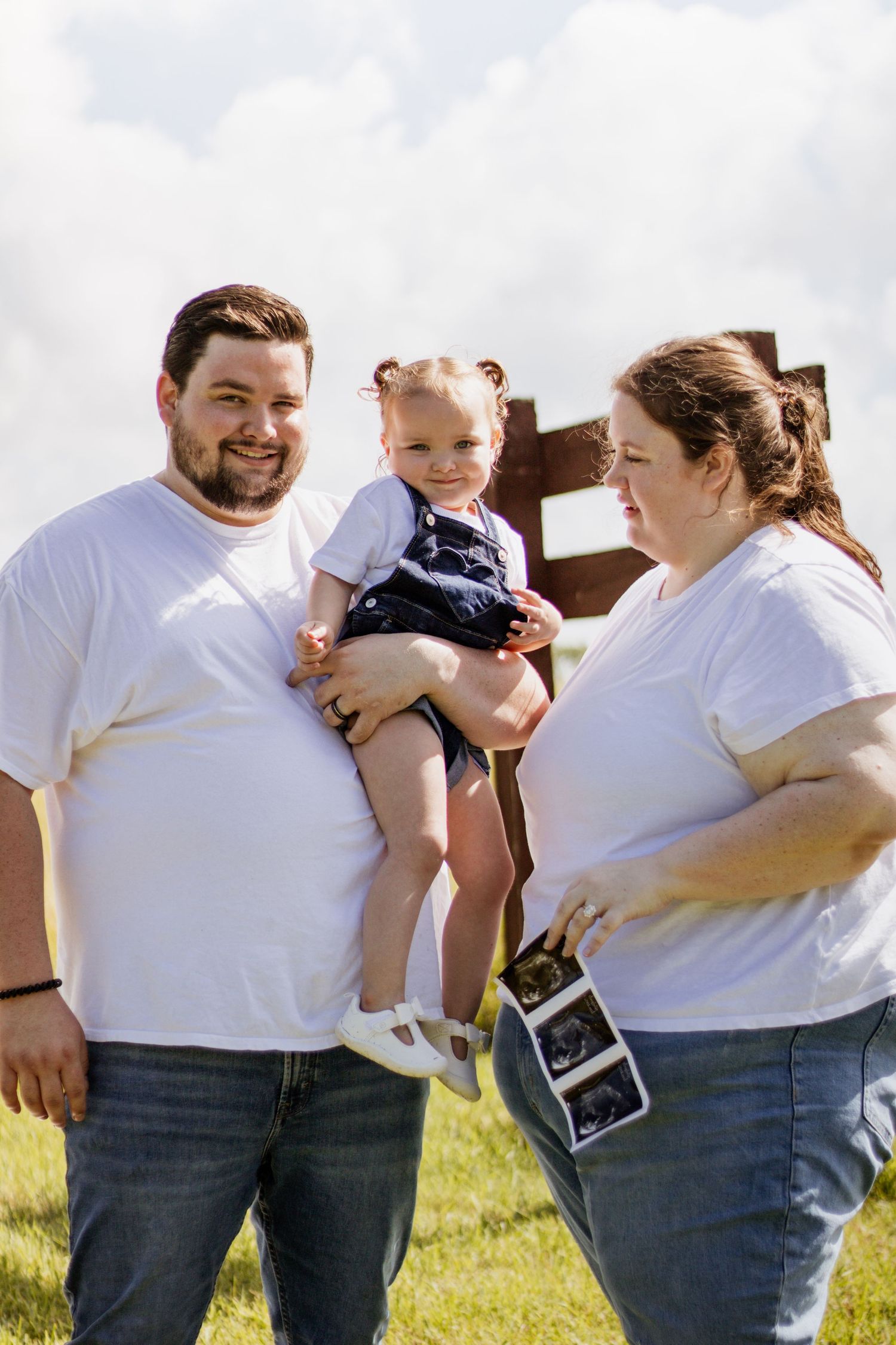 A couple in white t-shirts and jeans hold a toddler and ultrasound photos outdoors, announcing a pregnancy.