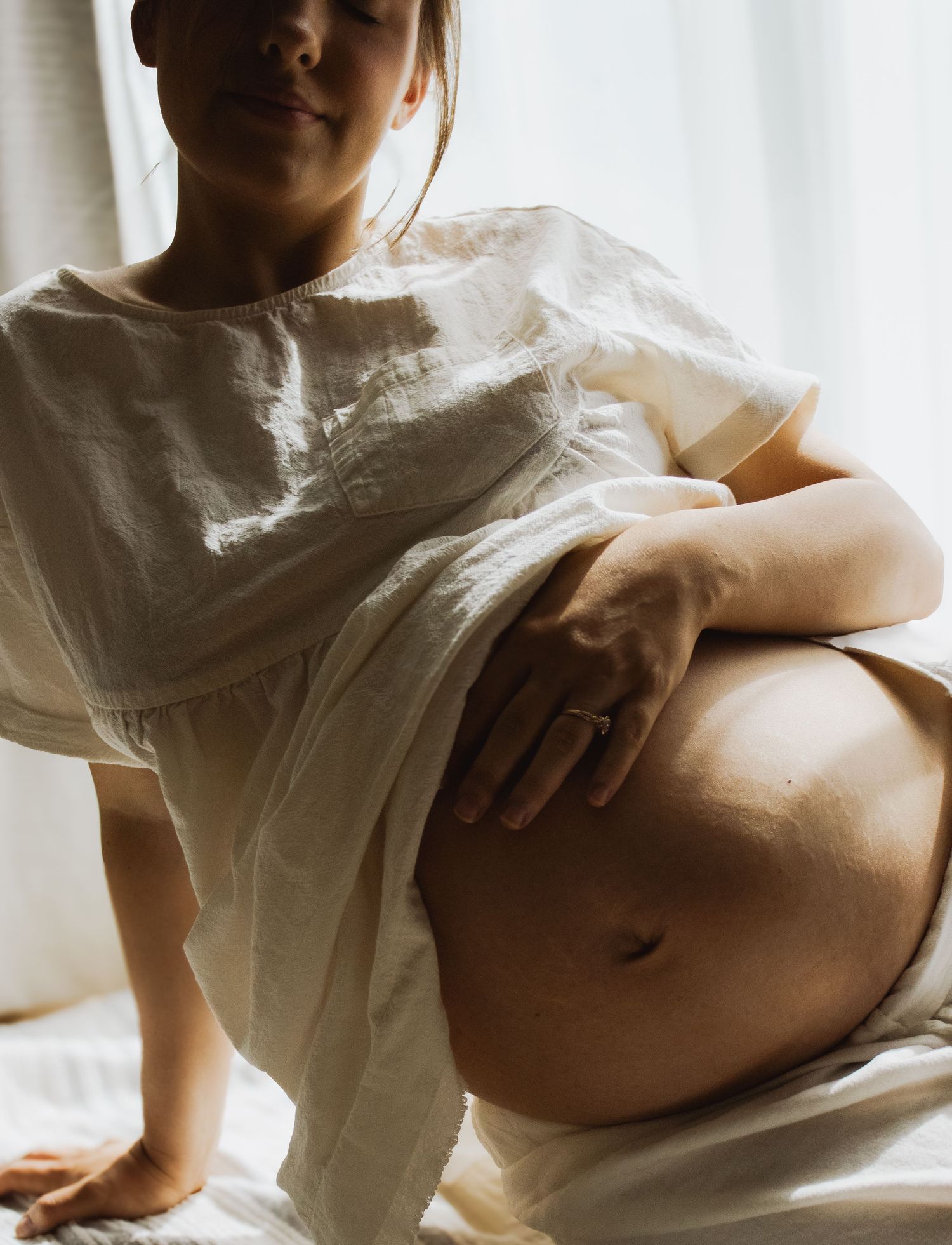 Pregnant woman in white linen top gently holding her large bare baby bump while sitting on white bed.