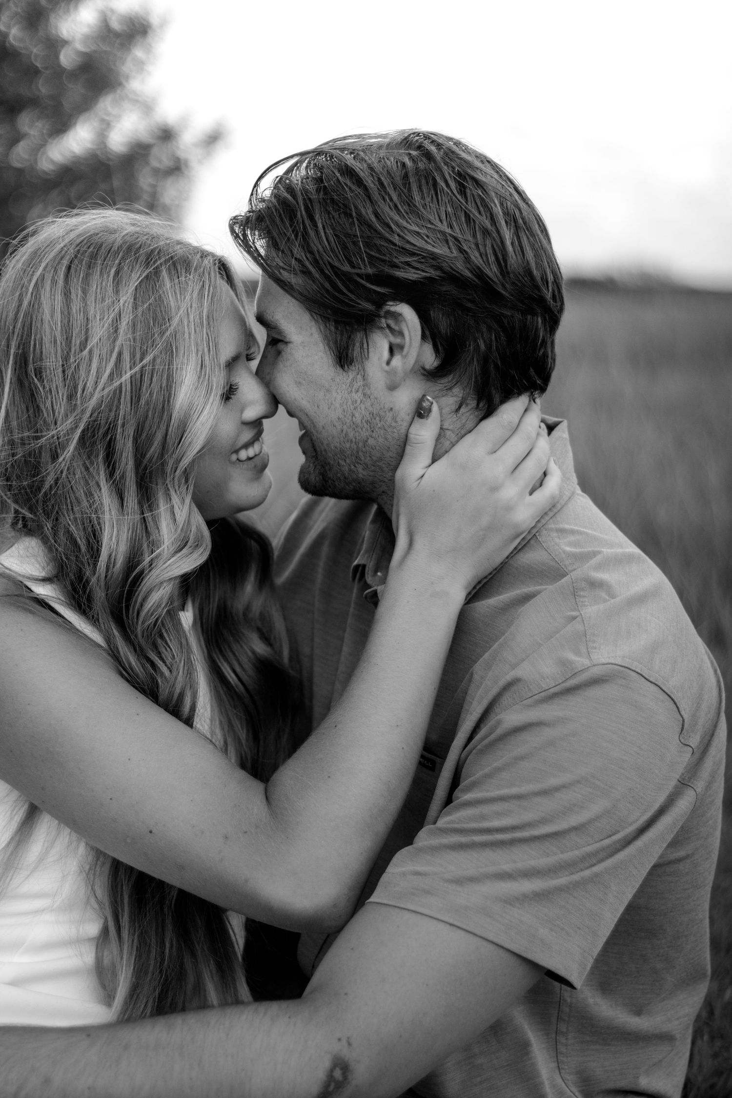 Black and white photo of a smiling couple sharing a romantic moment, woman holding man's face gently outdoors.