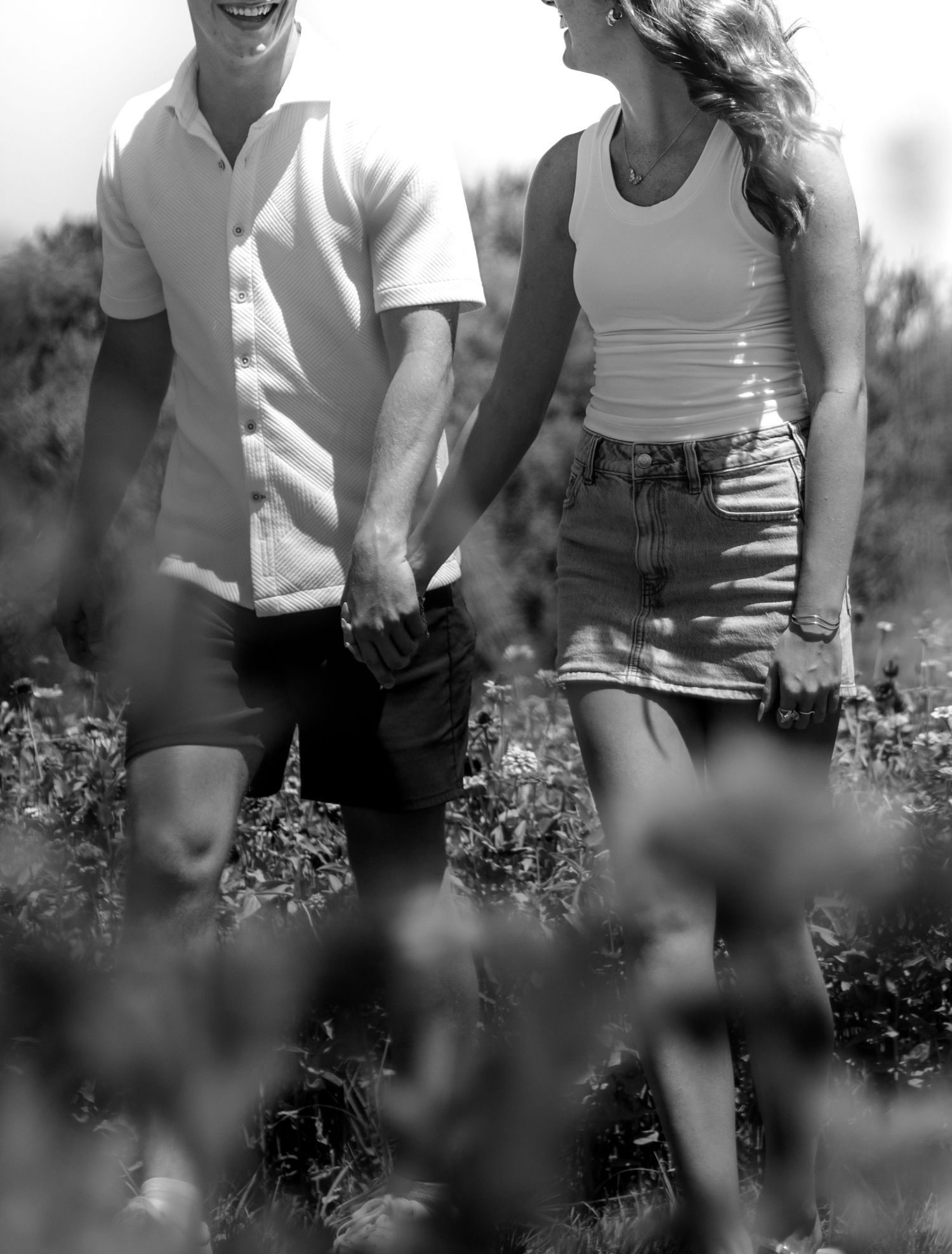 Black and white photo of a couple holding hands and walking through a wildflower field on a sunny day.