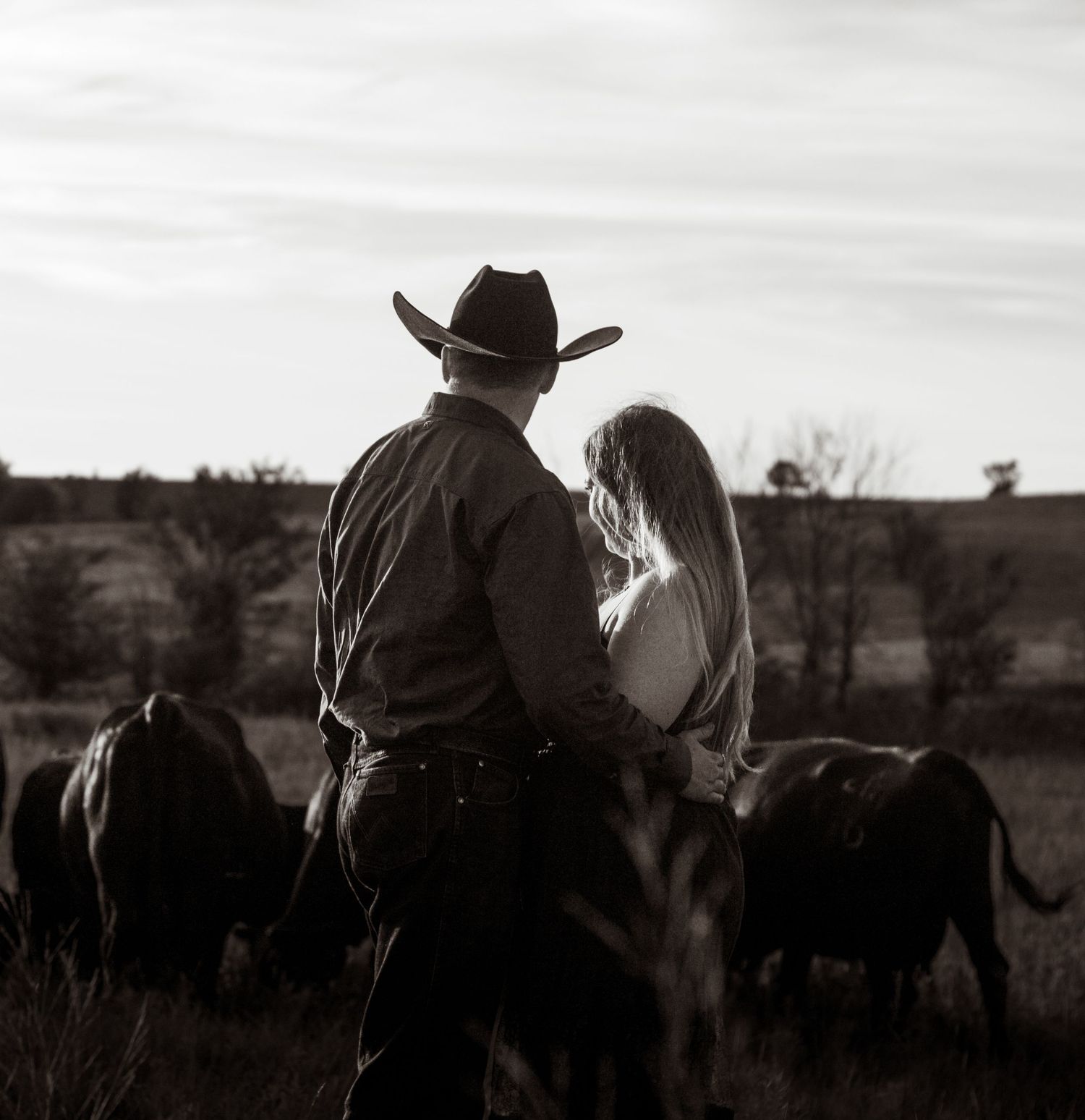 Black and white photo of a cowboy and woman standing together watching cattle graze on a rural ranch at dusk.