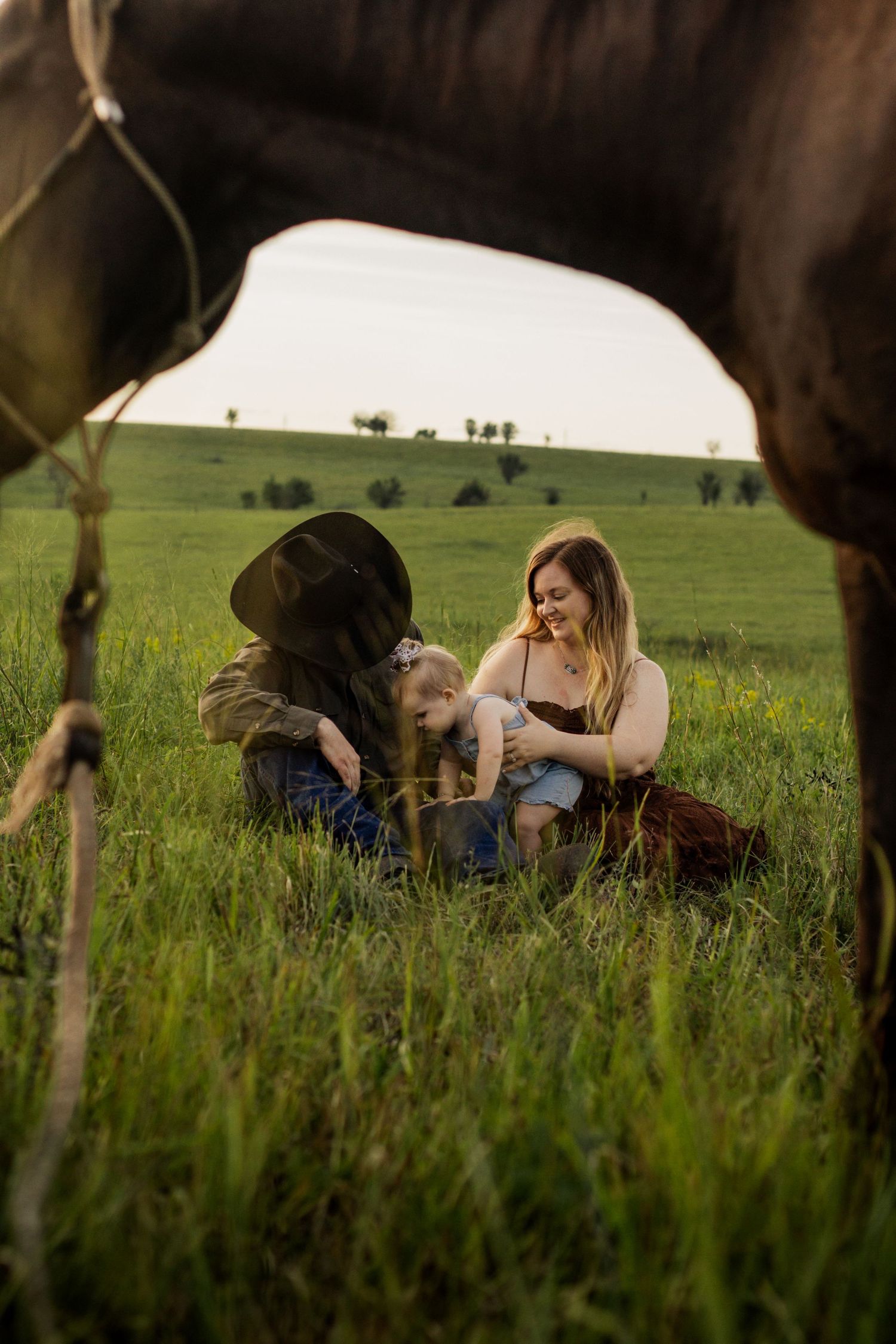 Two women and a child sit in a green field, framed by a horse, enjoying a warm golden hour moment.