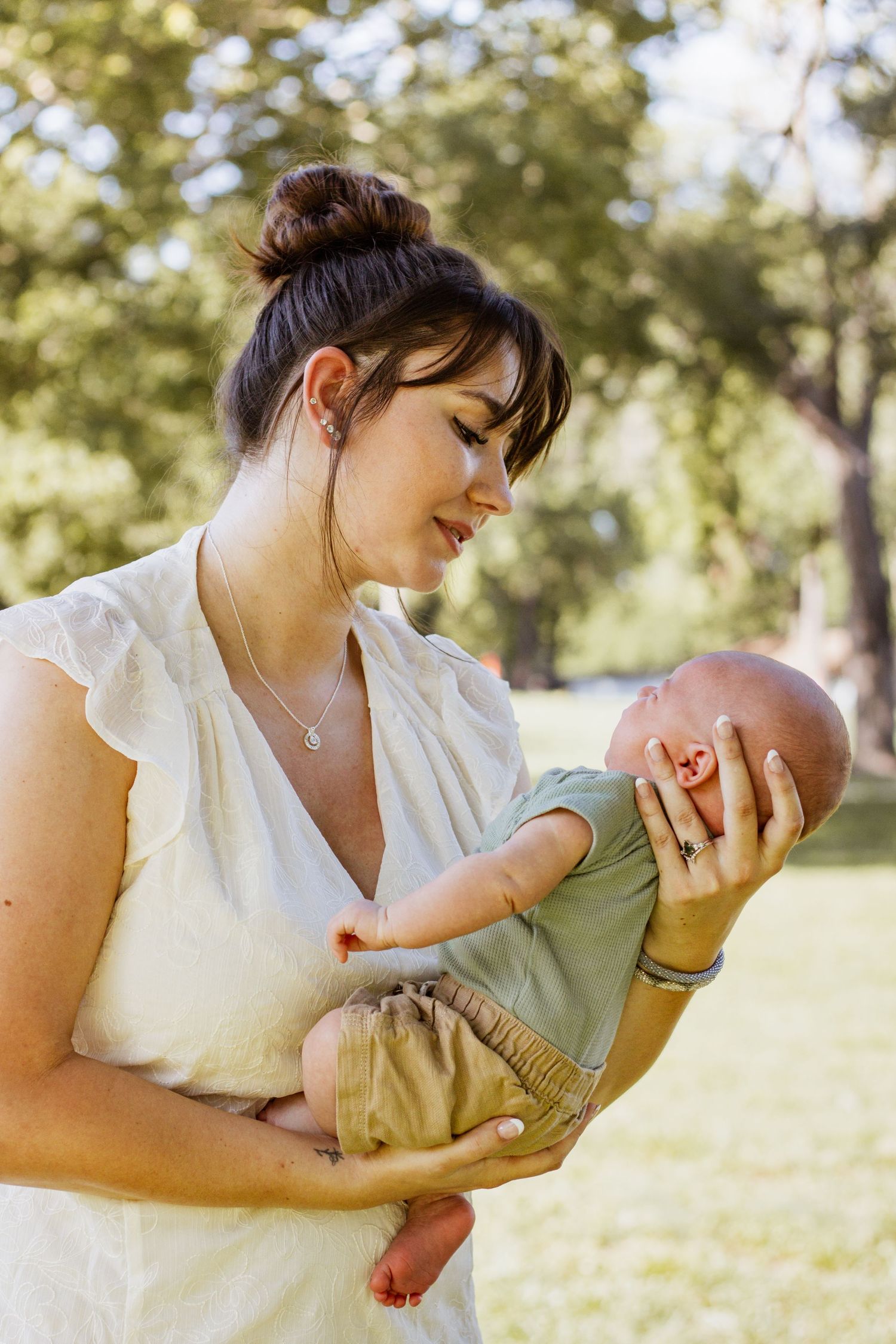 A young mother in a white dress tenderly holds her newborn baby outdoors in a sunny green park.