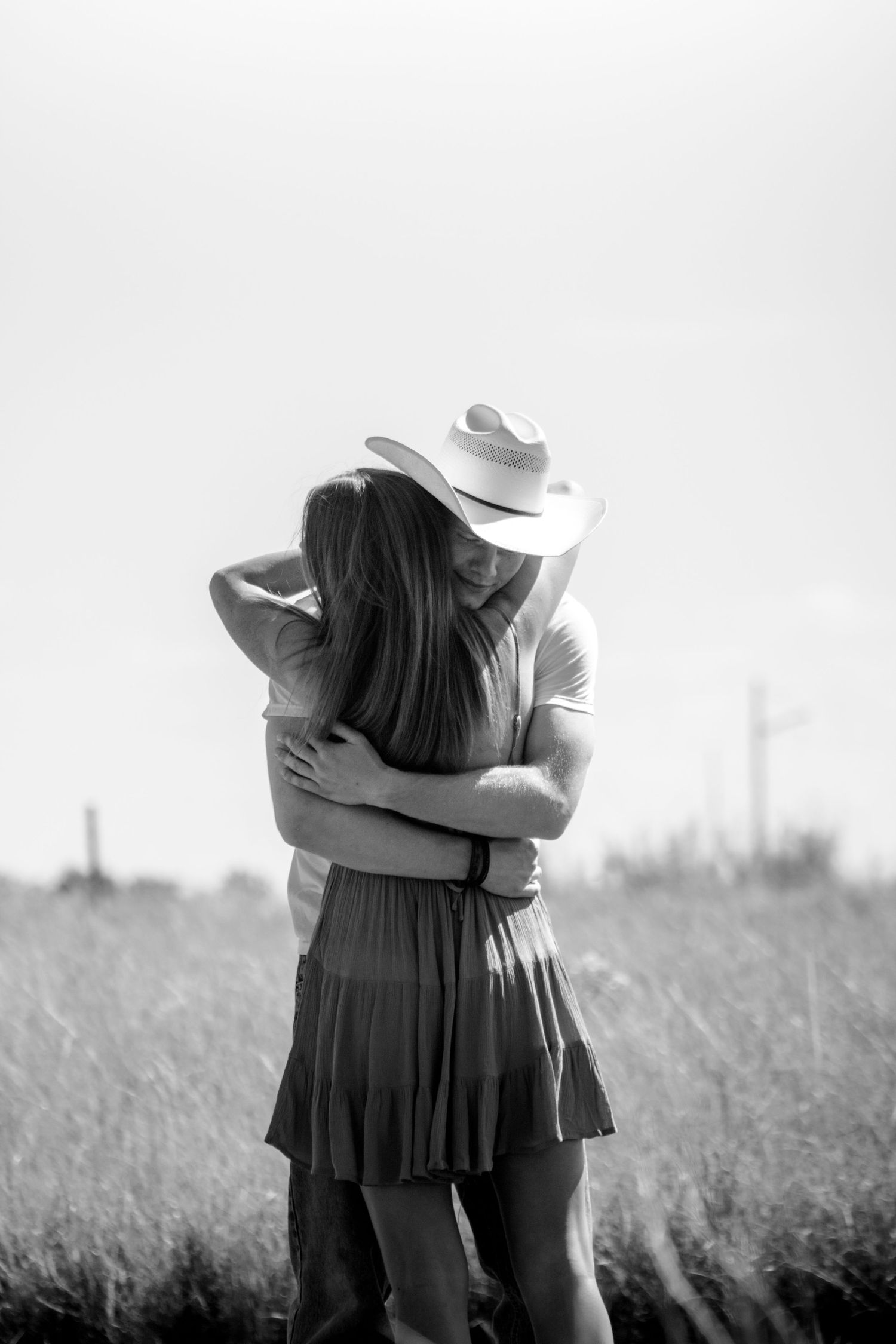 Black and white photo of a couple embracing in a field, man wearing a cowboy hat, romantic outdoor portrait.